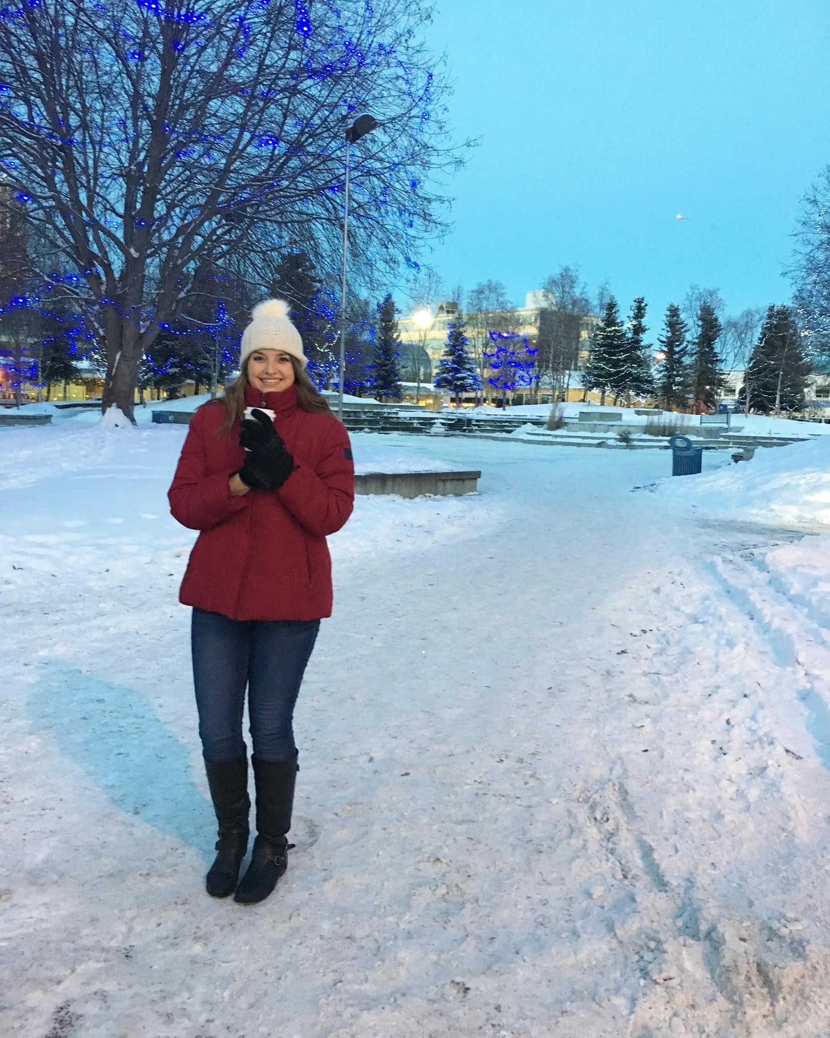 Leah wearing a red jacket and white hat outside in snow holding a hot drink.