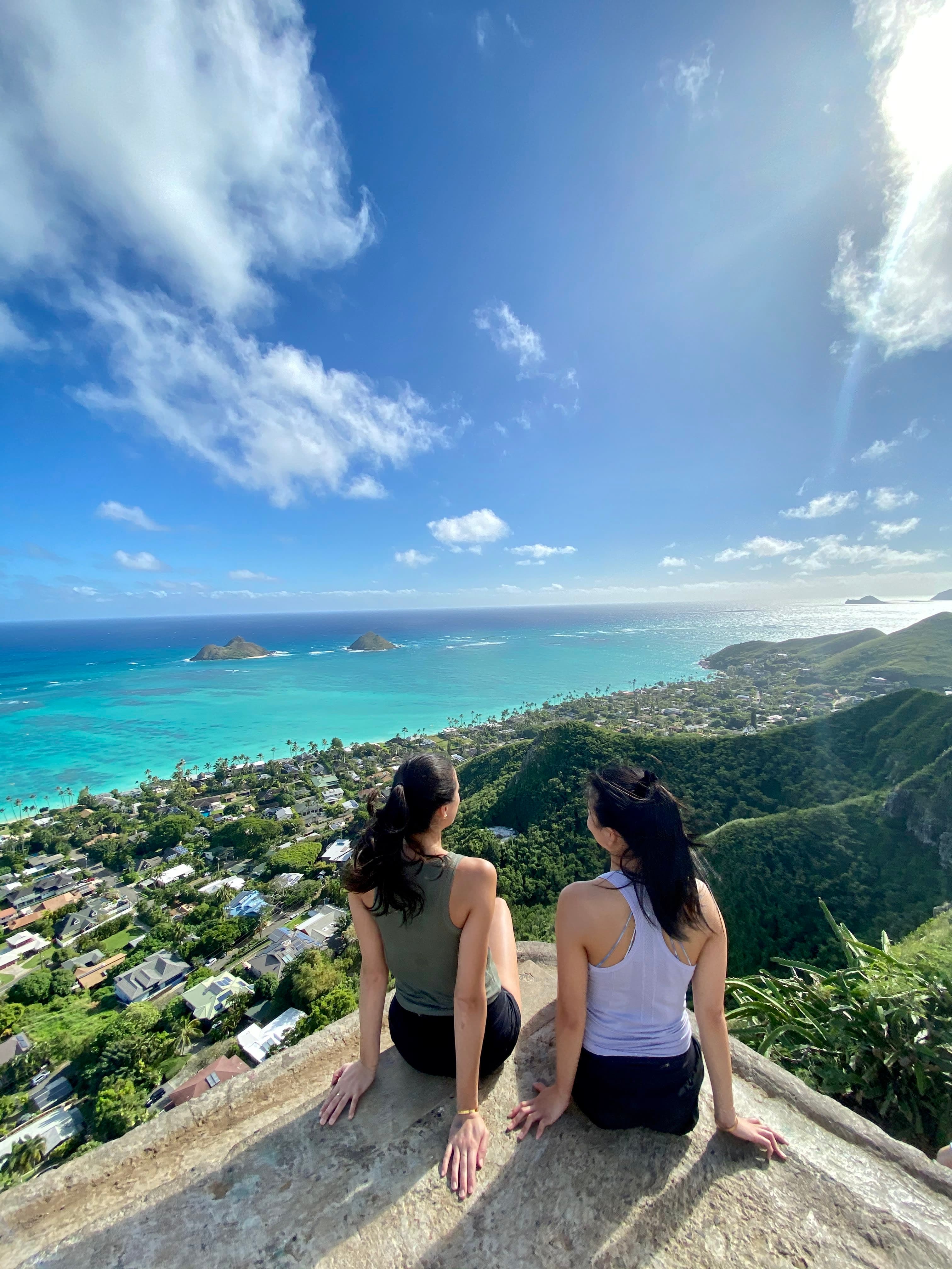 Michelle and a friend in hiking clothes sitting on a concrete platform overlooking green hills, buildings and the ocean.