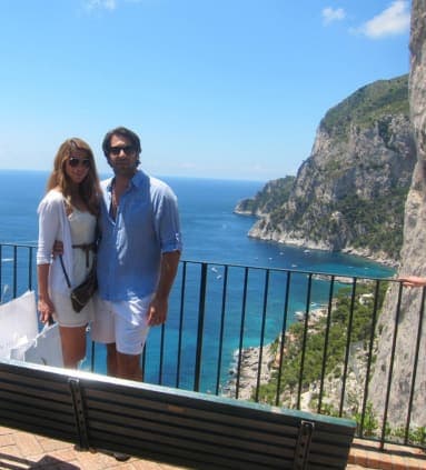 A man and woman posing on a balcony overlooking a coast line with cliffs and crystal clear blue water