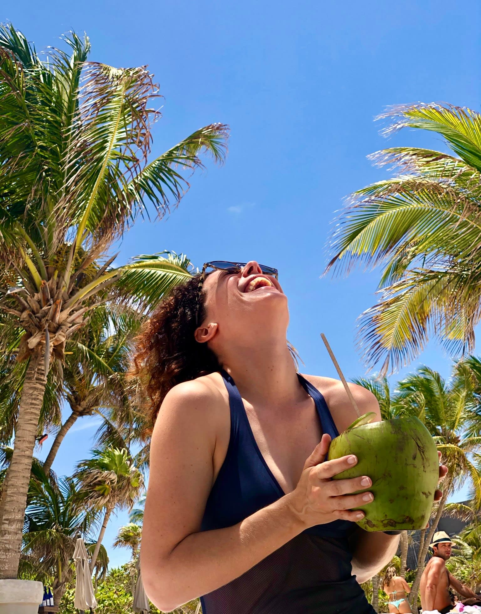 Travel advisor Alexandra looking up at the sky and smiling, holding a green coconut, with palm trees in view