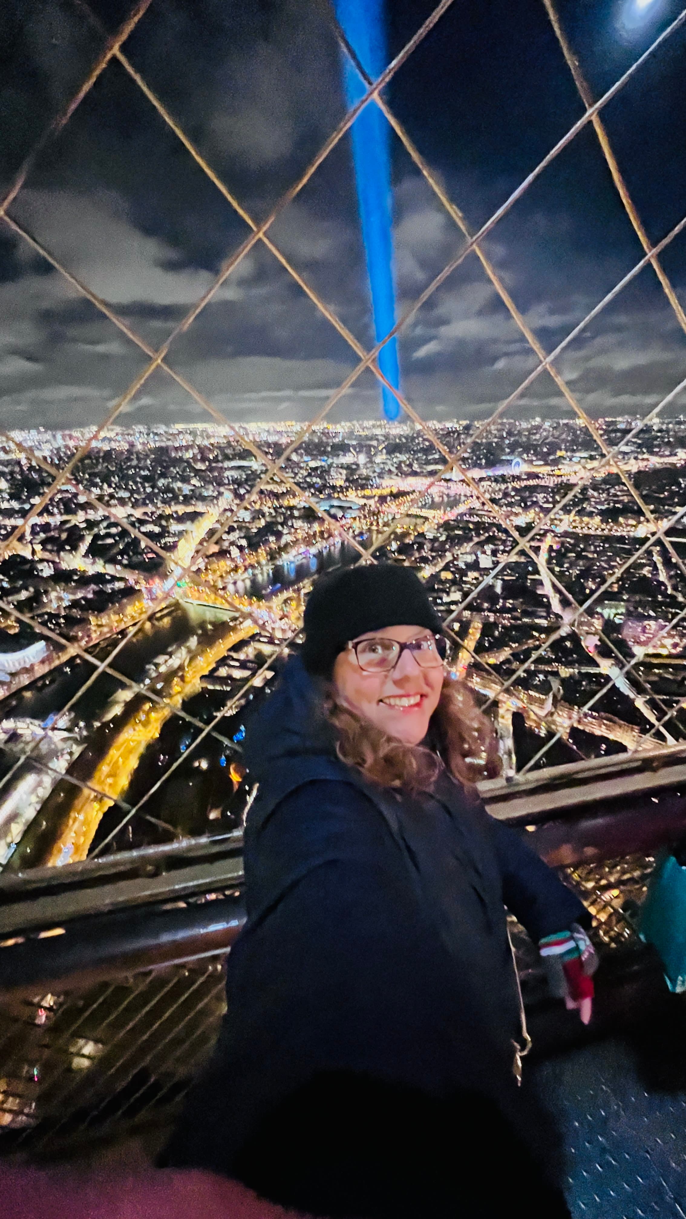 Vivian in a black jacket and beanie at the top of the Eiffel Tower with Paris in the background