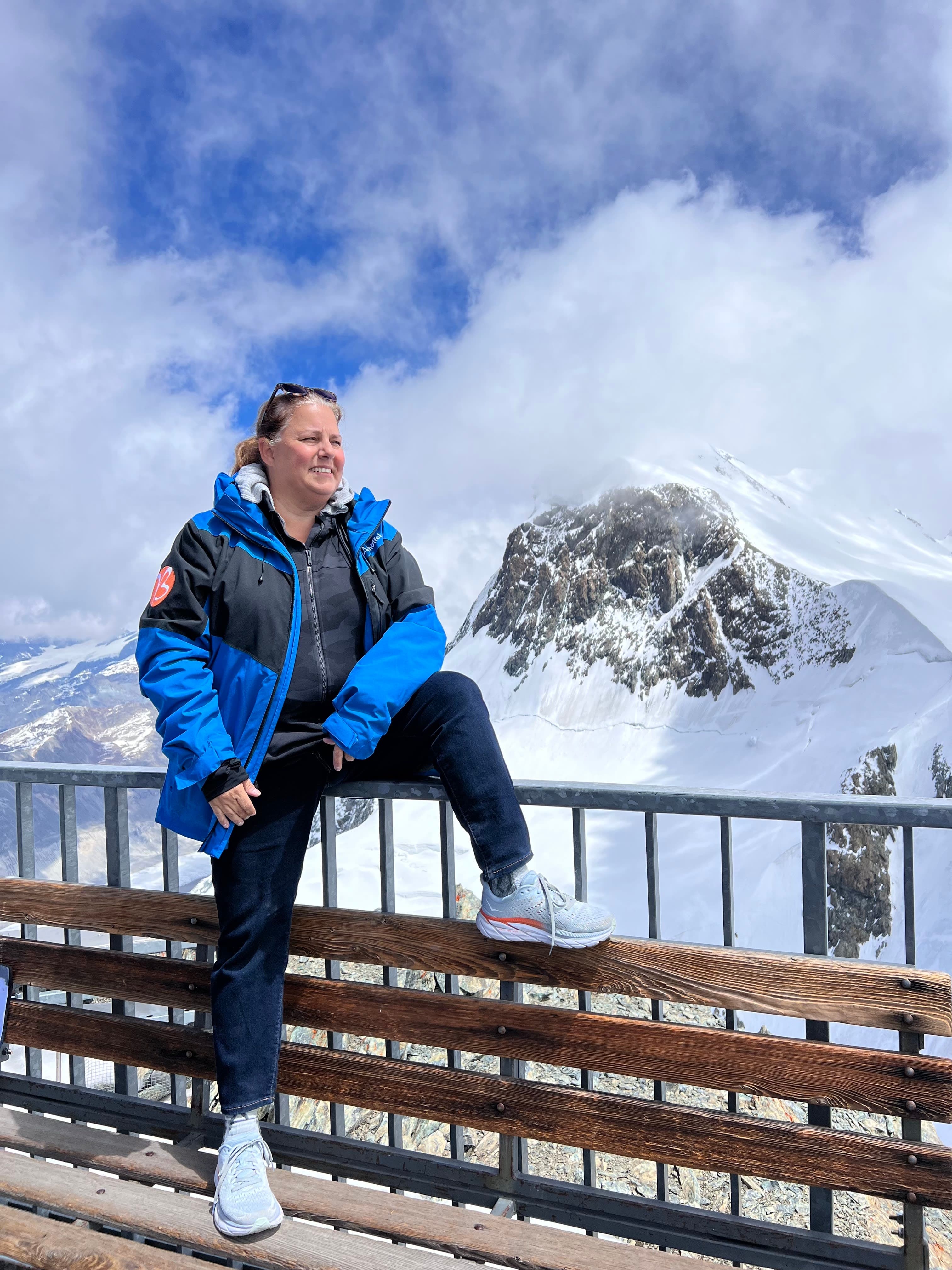 Vivian in a blue jacket posing for a photo on a terrace with snow capped mountains in the background.