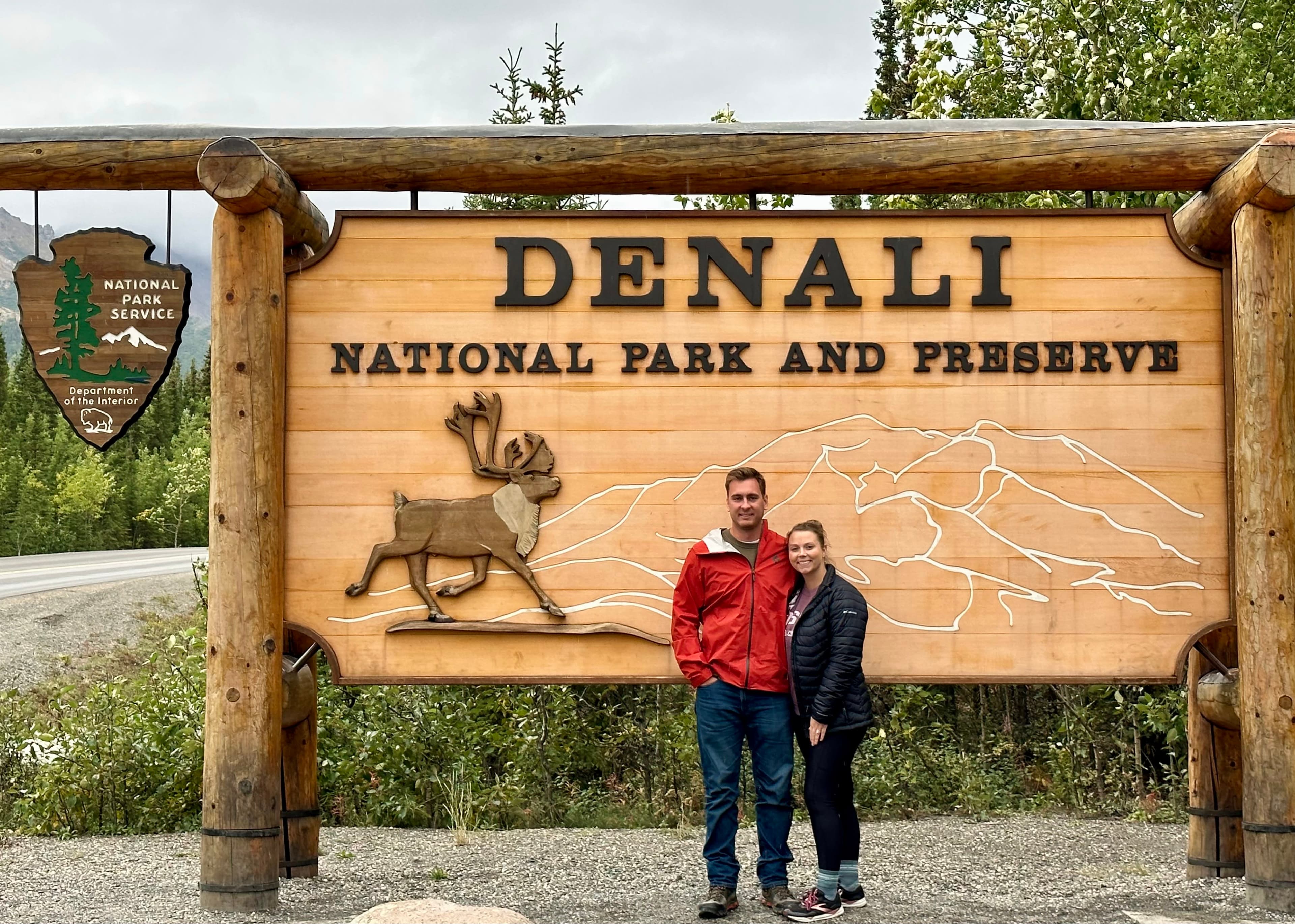 Elizabeth and a man standing in front of the wooden Denali National Park and Preserve sign.