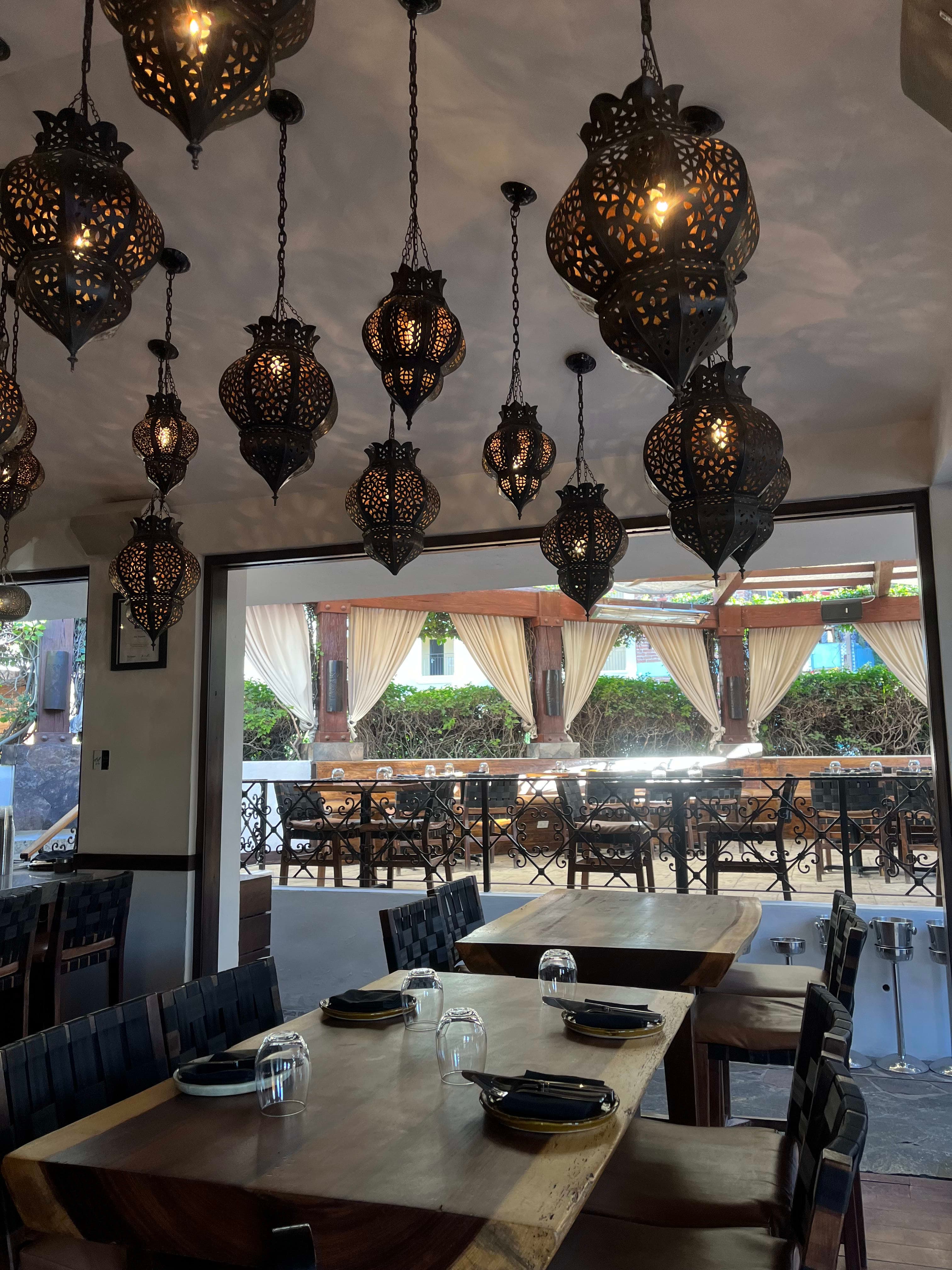 An open-air indoor restaurant with wooden tables and antique lamps hanging from the ceiling, with a covered patio beyond.