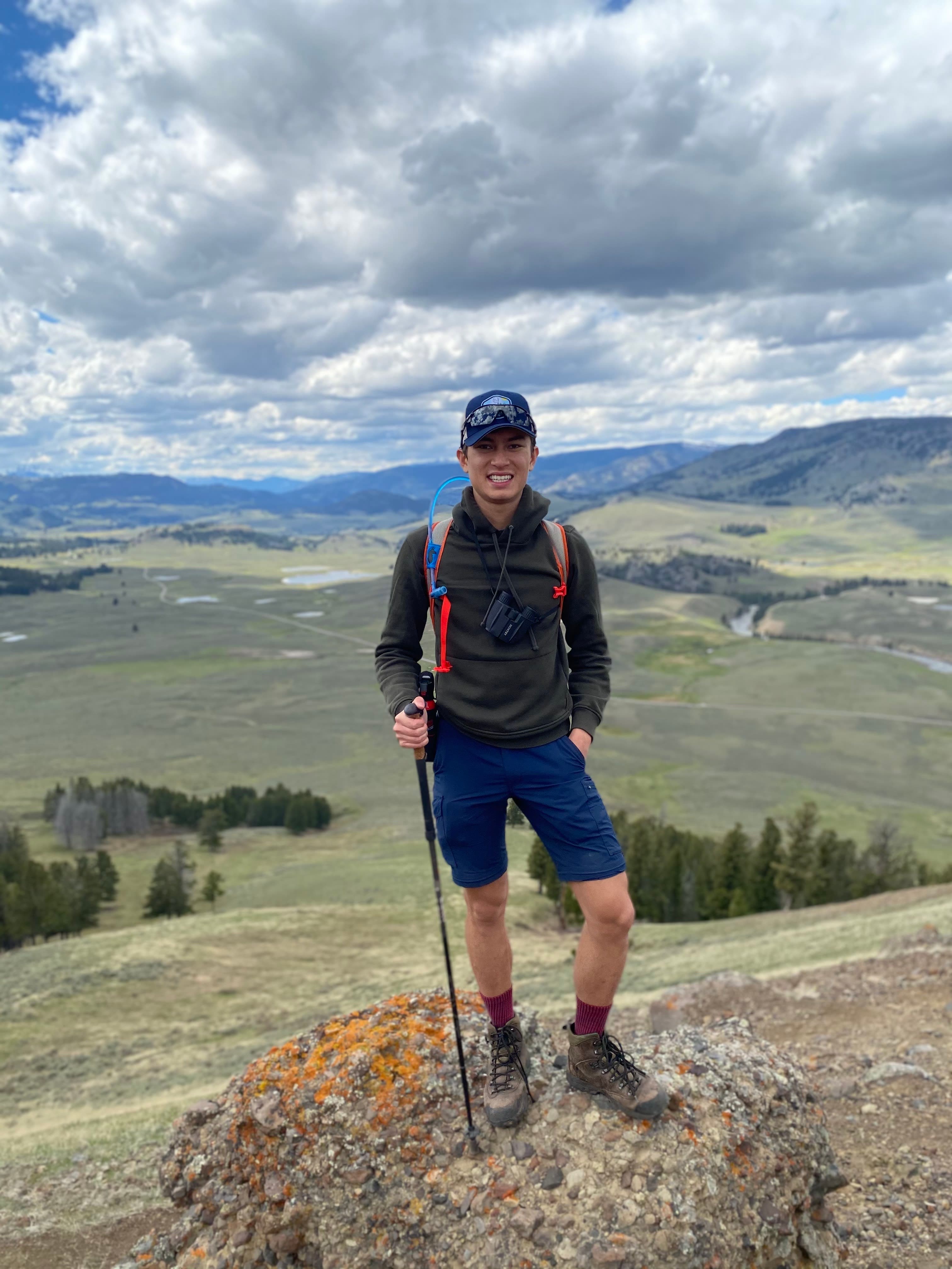 Justin in hiking gear with a trekking pole standing on a rock overlooking a green valley on a nice day with clouds