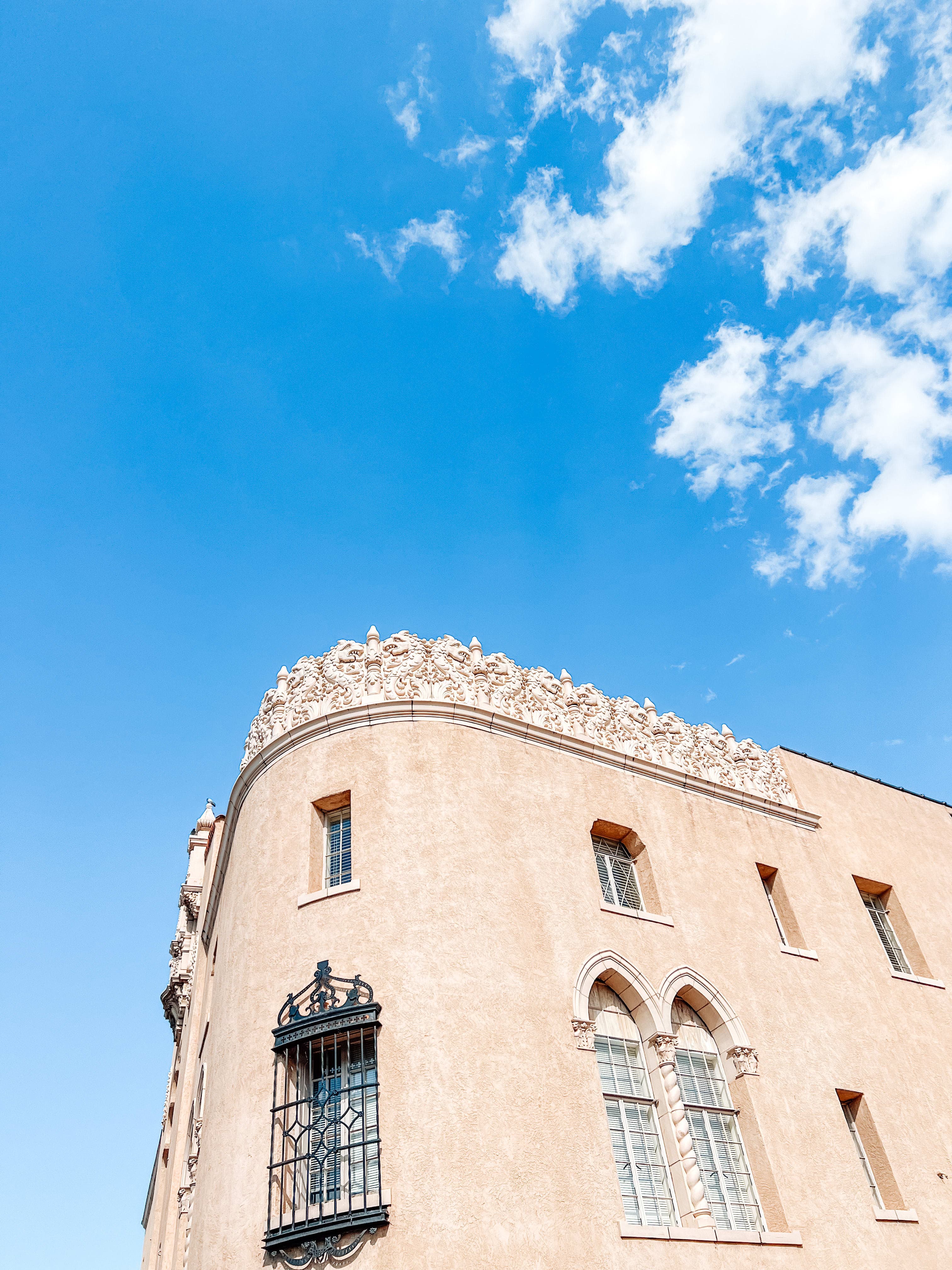 A photo of a sand-colored, corner building from below with a blue sky and clouds in the background.