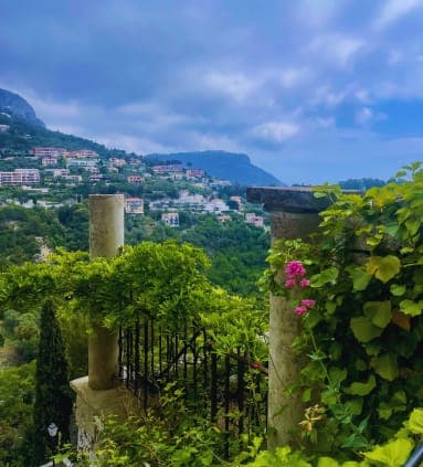 View of a small town on a cliffside from a balcony, featuring a cloudy sky, outdoor plants, trees, mountains, grass, flowers, and a garden.
