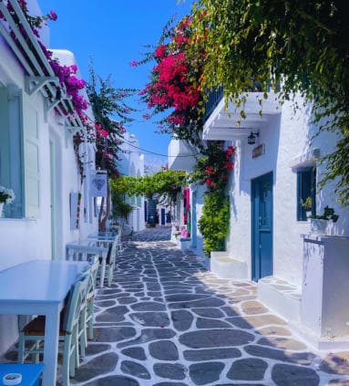 View of a stone street in Paros, Greece, with tables and chairs set up outdoors, it includes plants, flowers, and a clear blue sky.