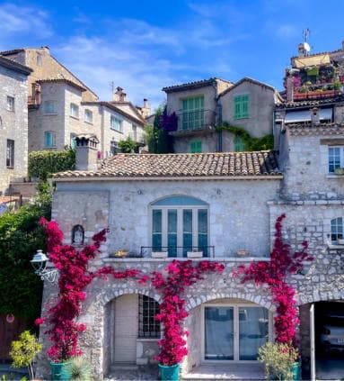 View of a stone building in St. Paul de Vence, France, a blue sky, and flowers in flowerpots on the front.