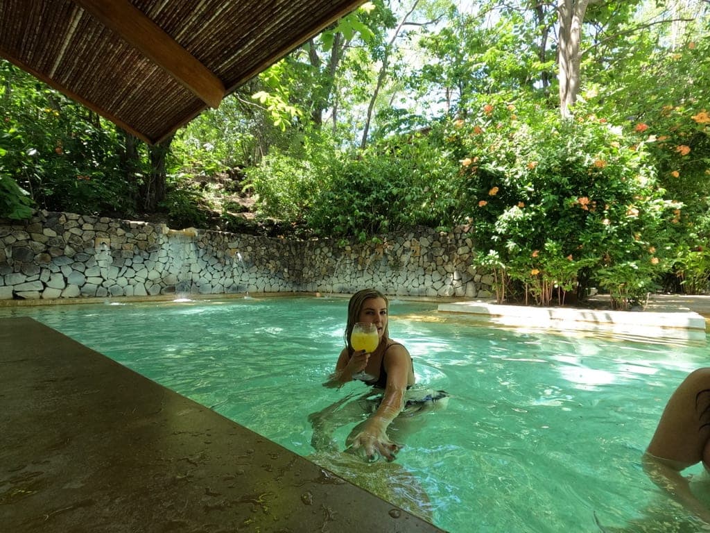 Stefanie swimming in a pool surrounded by plants and trees with a drink in her hand