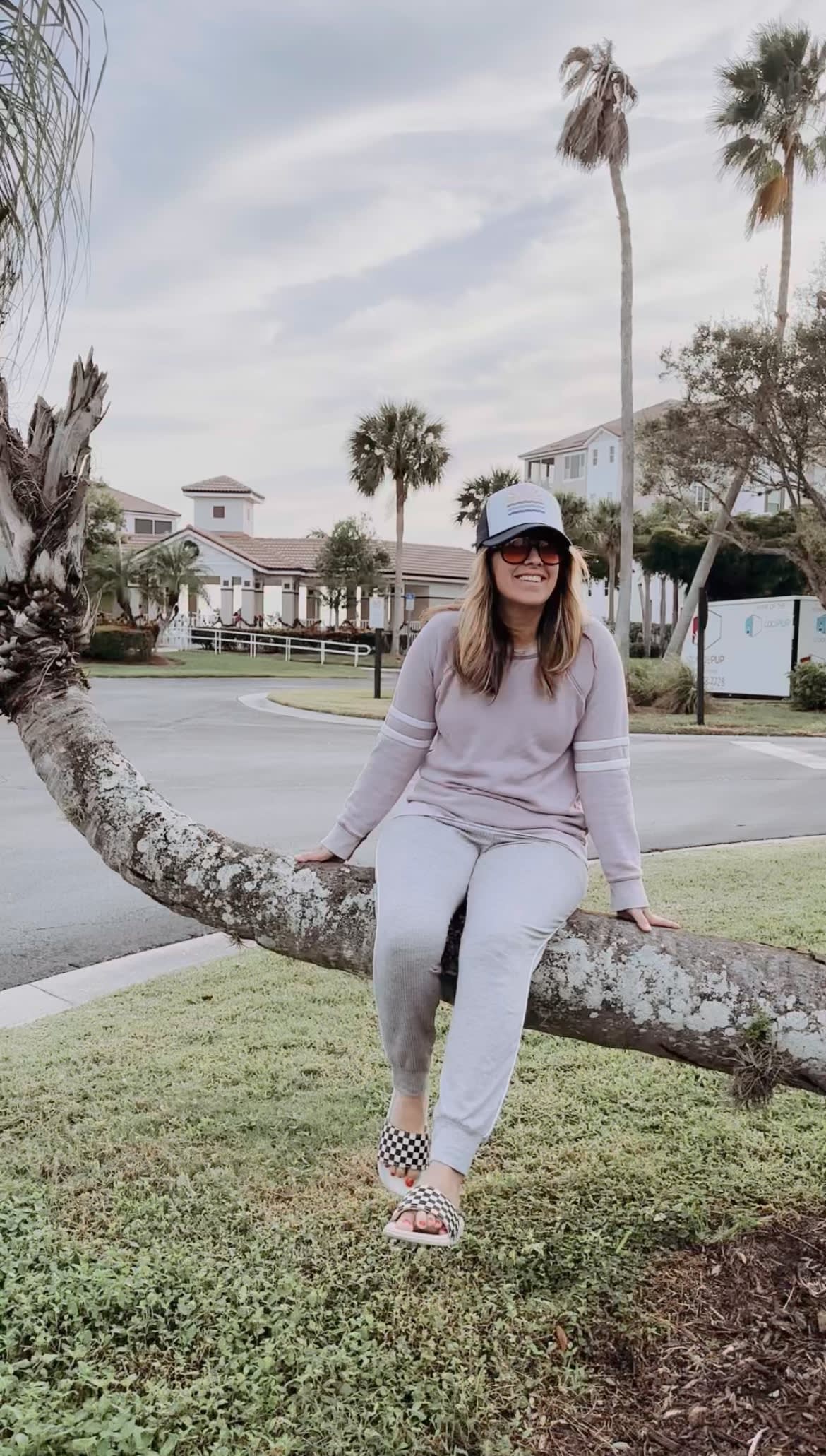 Picture of Leslie sitting on a low hanging palm tree branch in sweatpants, a hat and sunglasses.