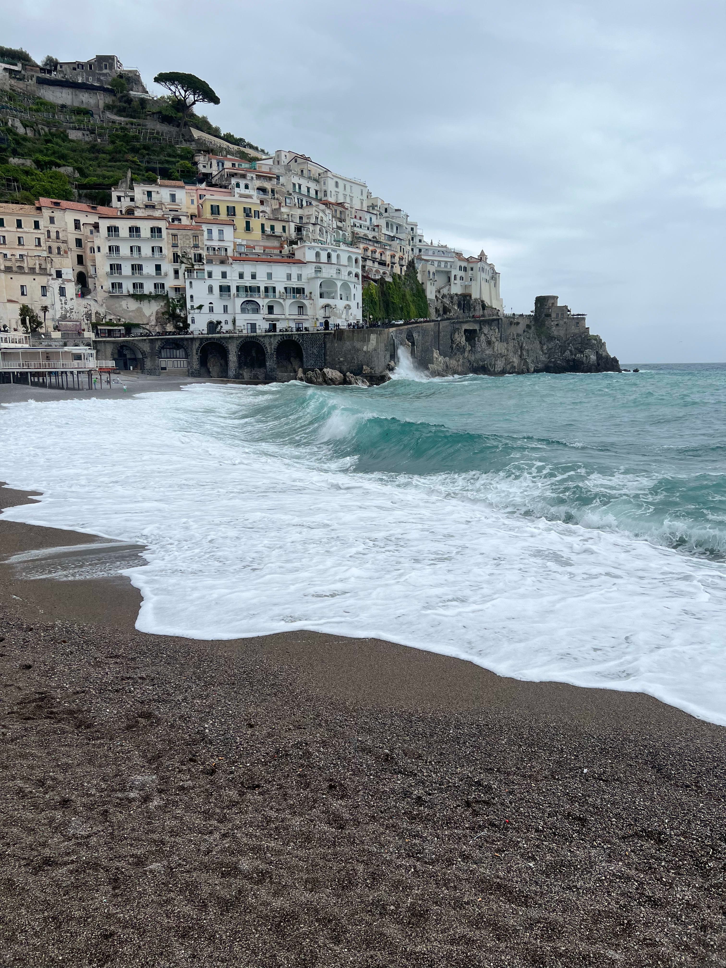 View of white sea foam waves crashing on the beach, buildings of the Amalfi Coast are also visible in the background
