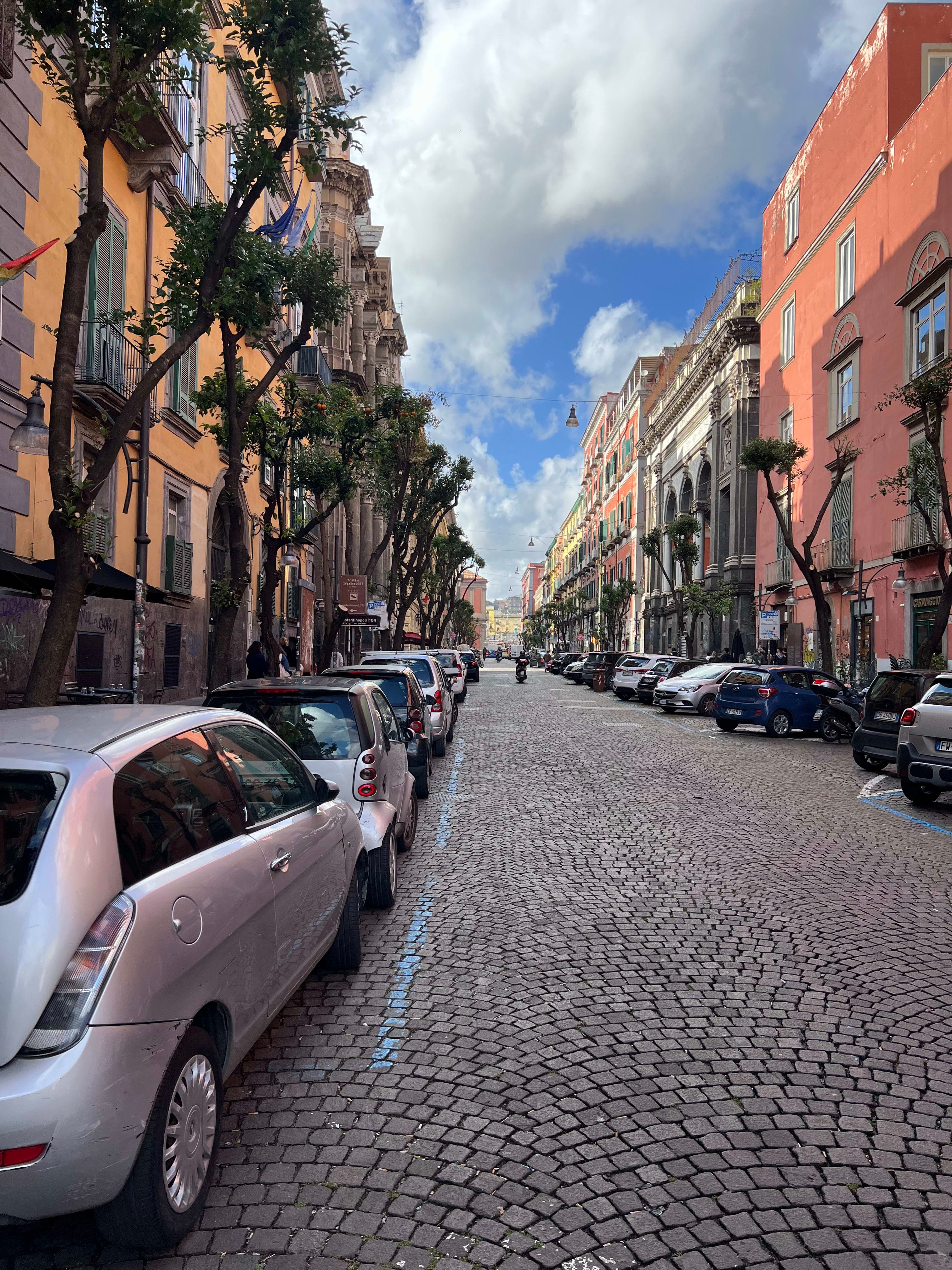 A cobblestone street in Naples with cars parked along the curb, on a cloudy day.