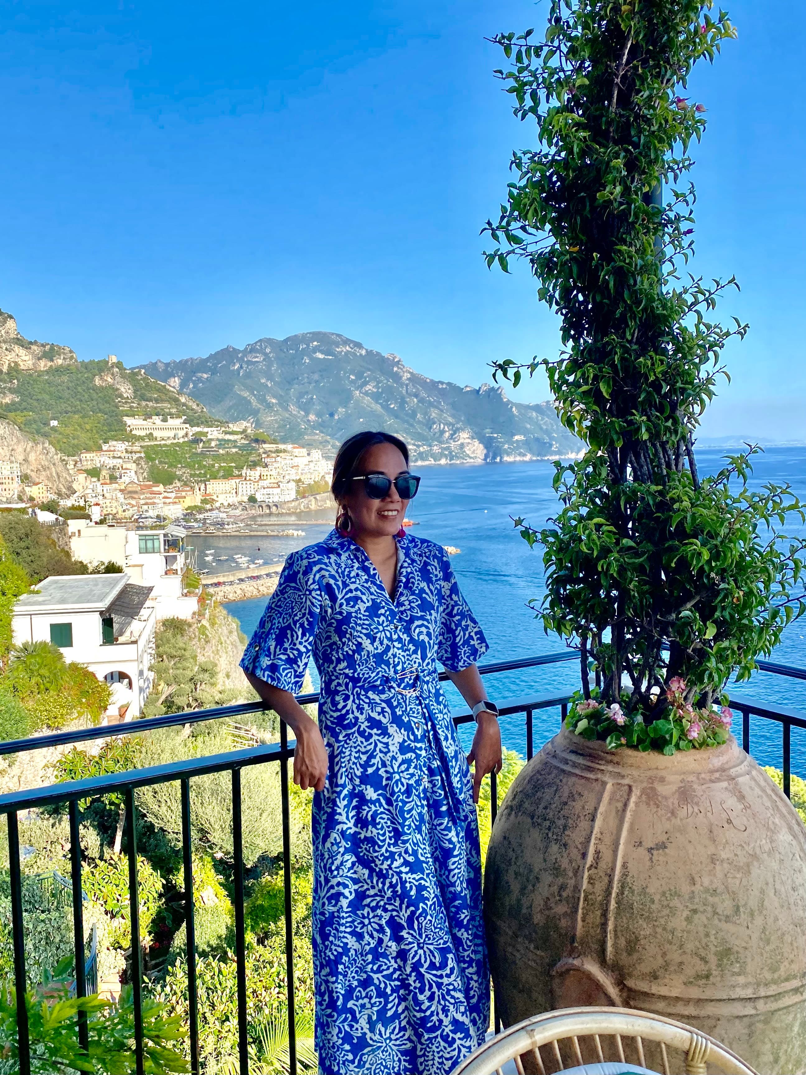Pam standing on a balcony in a blue dress with beautiful view of the sea and the Amalfi Coast in the background