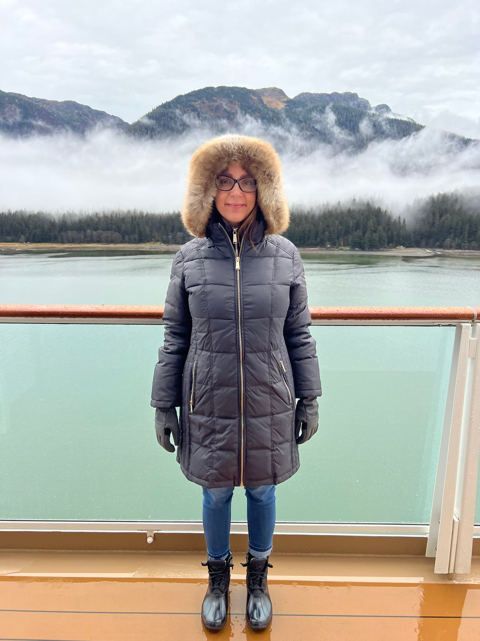 Megan in a parka standing on a cruise ship in front of turquoise water and mountains.