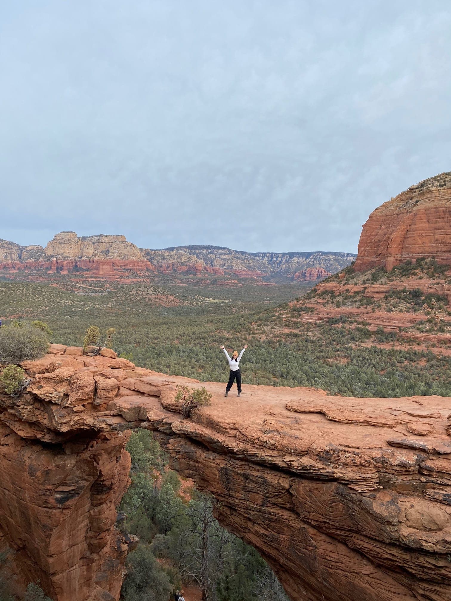 Isabella standing on a natural red rock bridge with her arms in the air with more red rocks in the background.