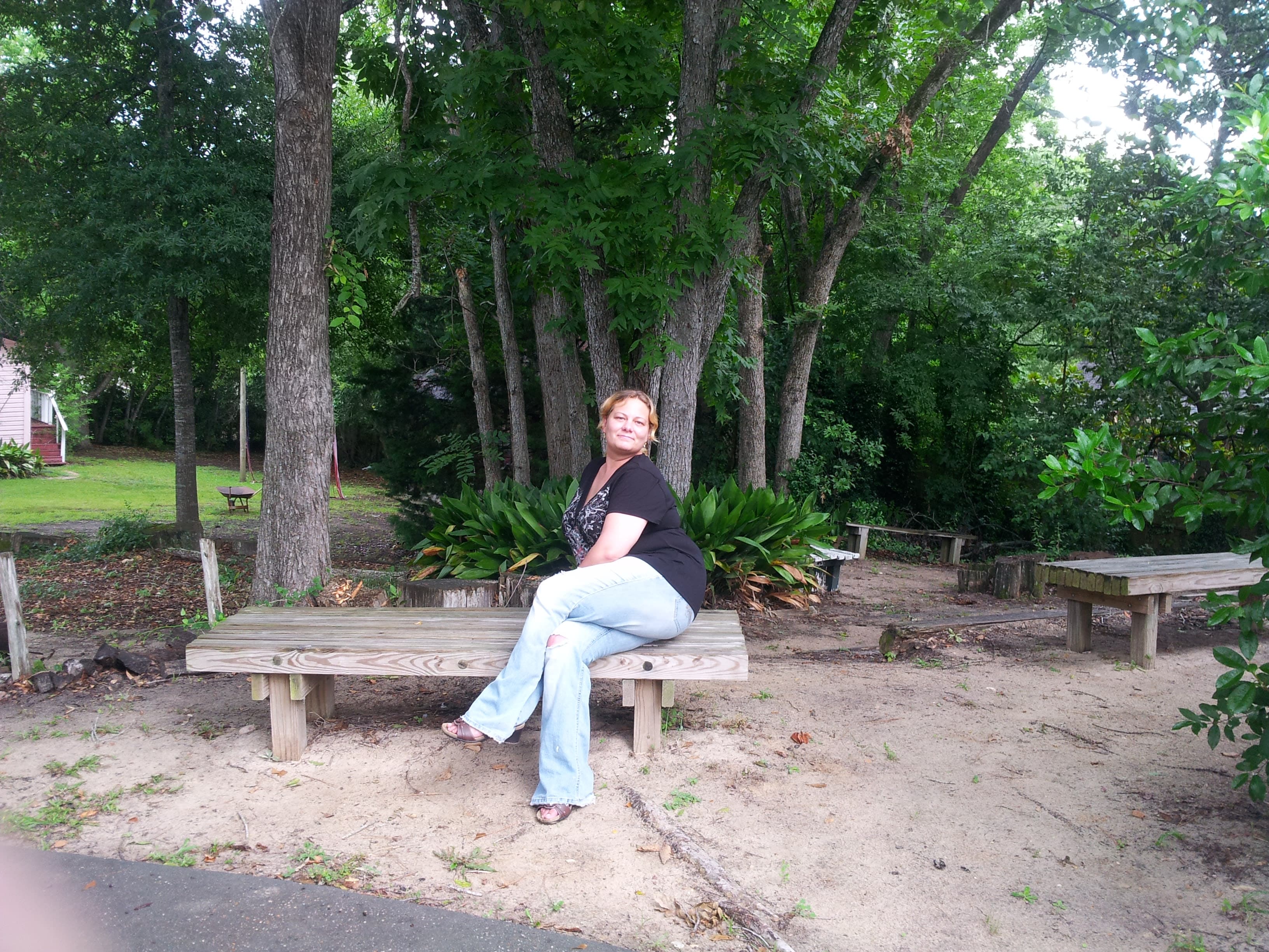 Picture of Wendy with her legs crossed wearing jeans and posing on a wooden bench in front of trees