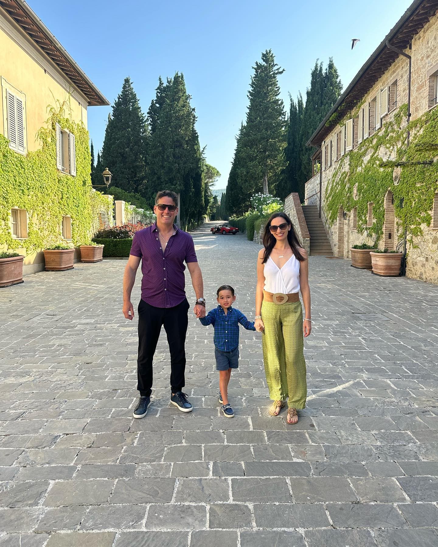 Picture of Mindy in green pants holding hands with family on a stone road in between two rows of buildings covered in vines