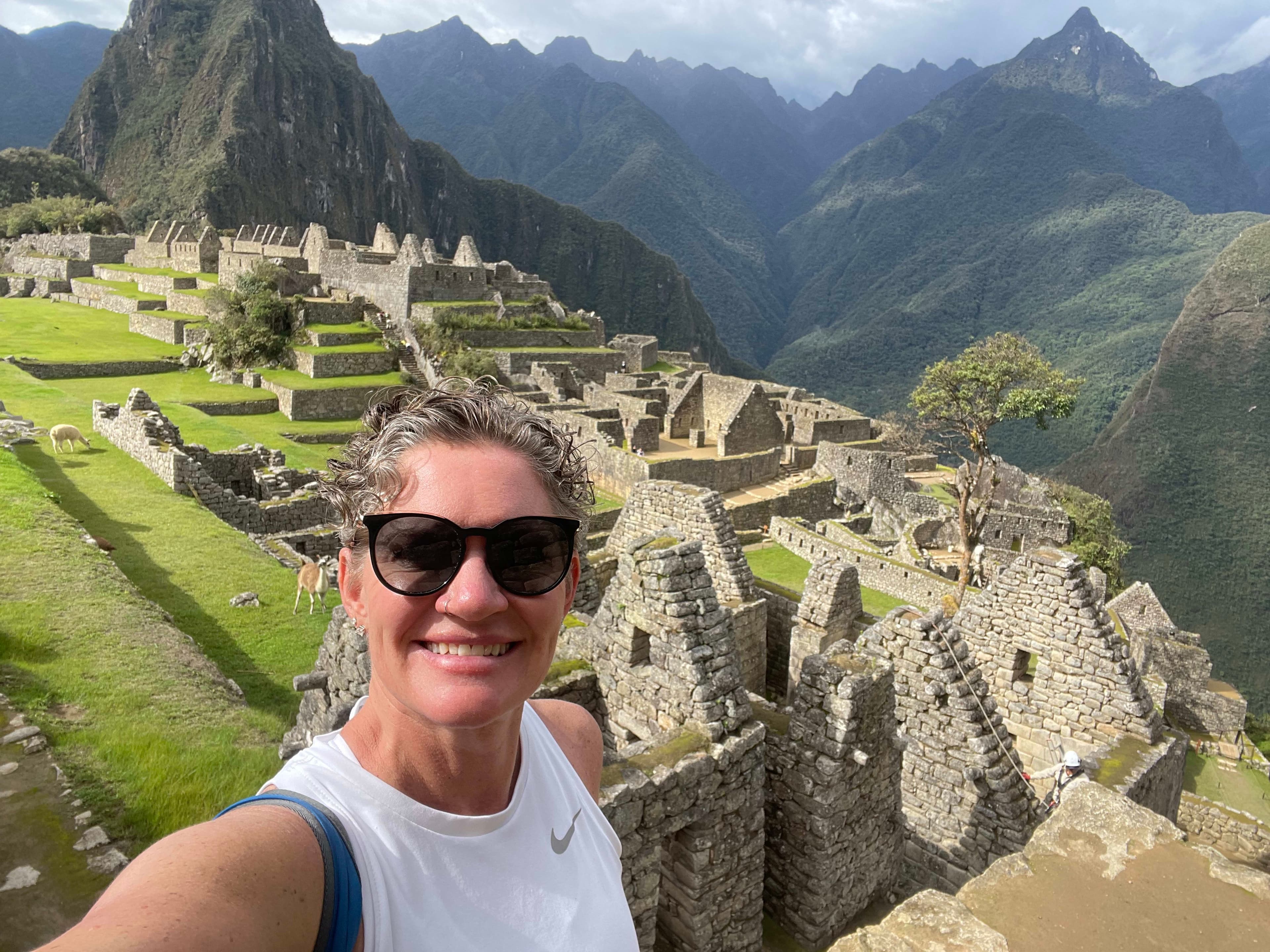 Selfie of Dayna wearing a white tank top and sunglasses with the Machu Picchu ruins in the background