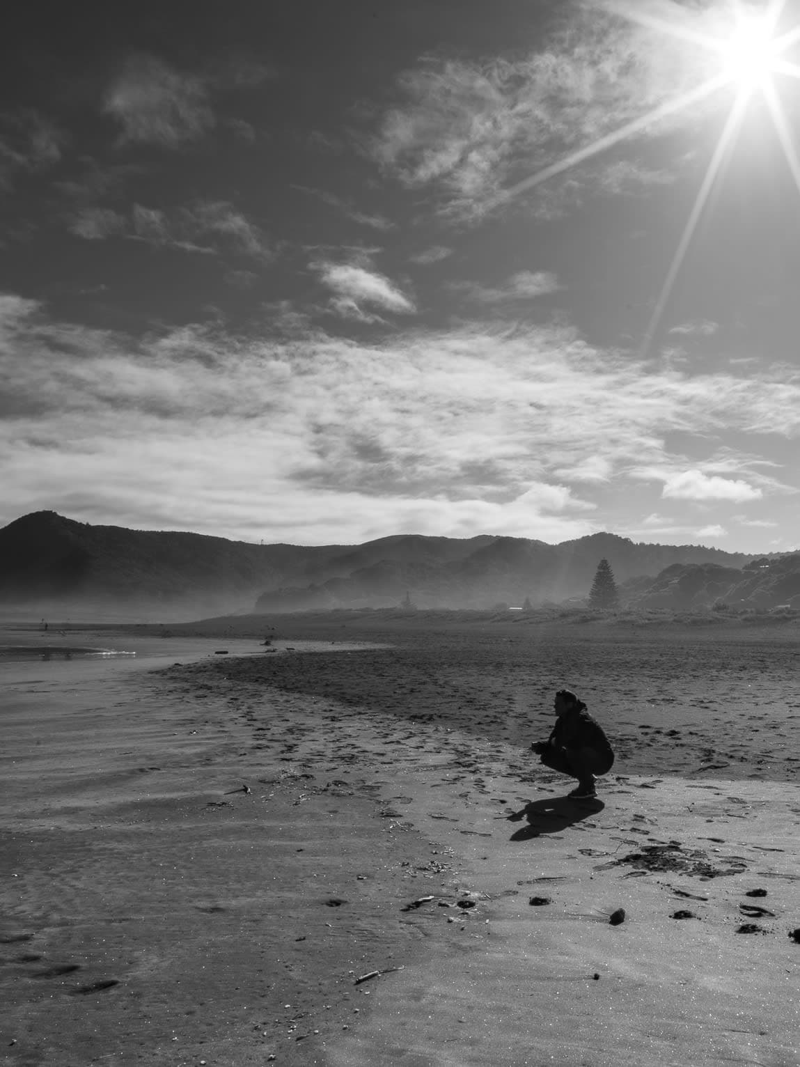 A grey scale image of Andrew sitting on a beach with sun shining bright in the corner
