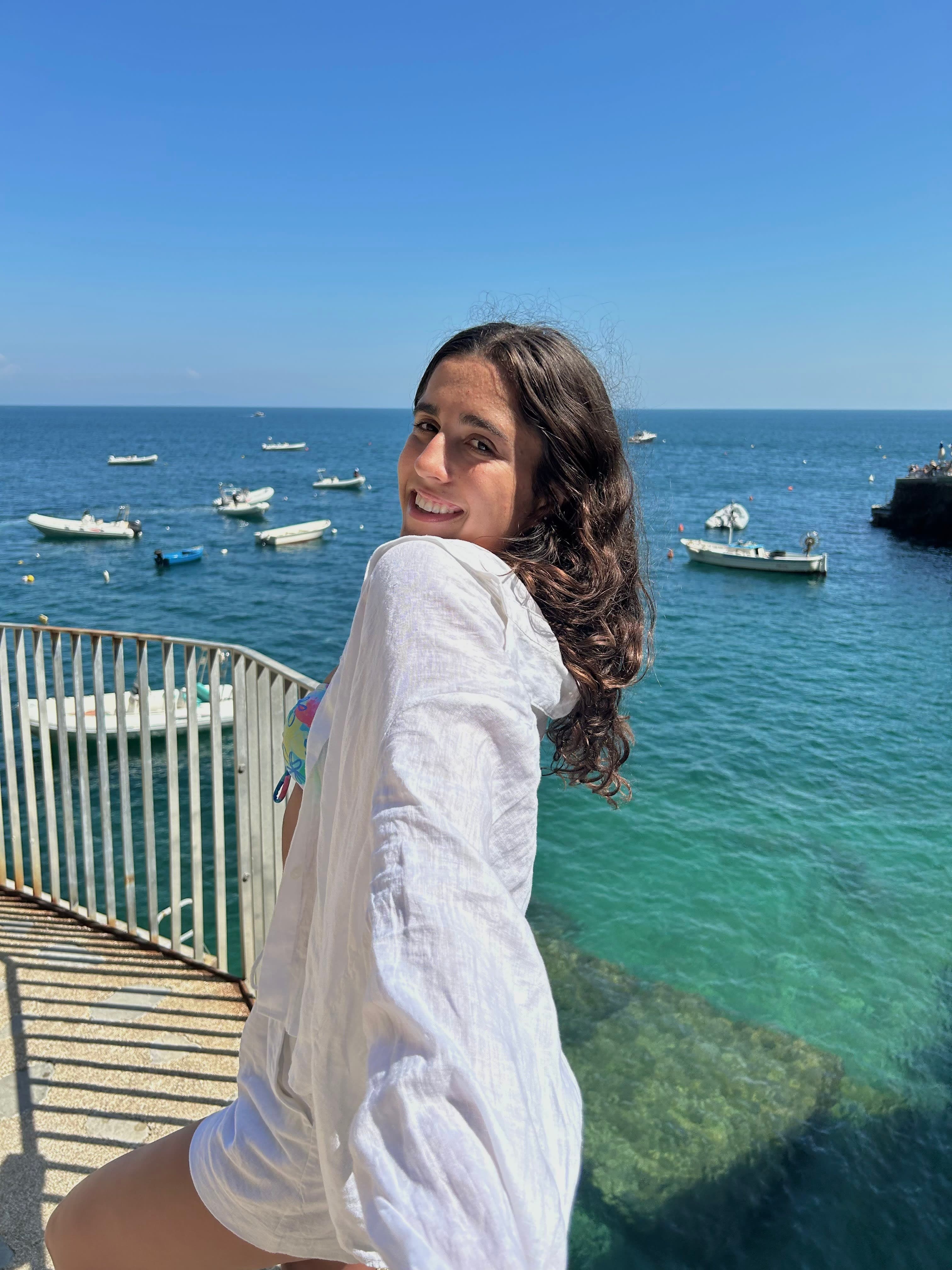 Avery standing on the deck in a white shirt with the view of the sea and boats in the background