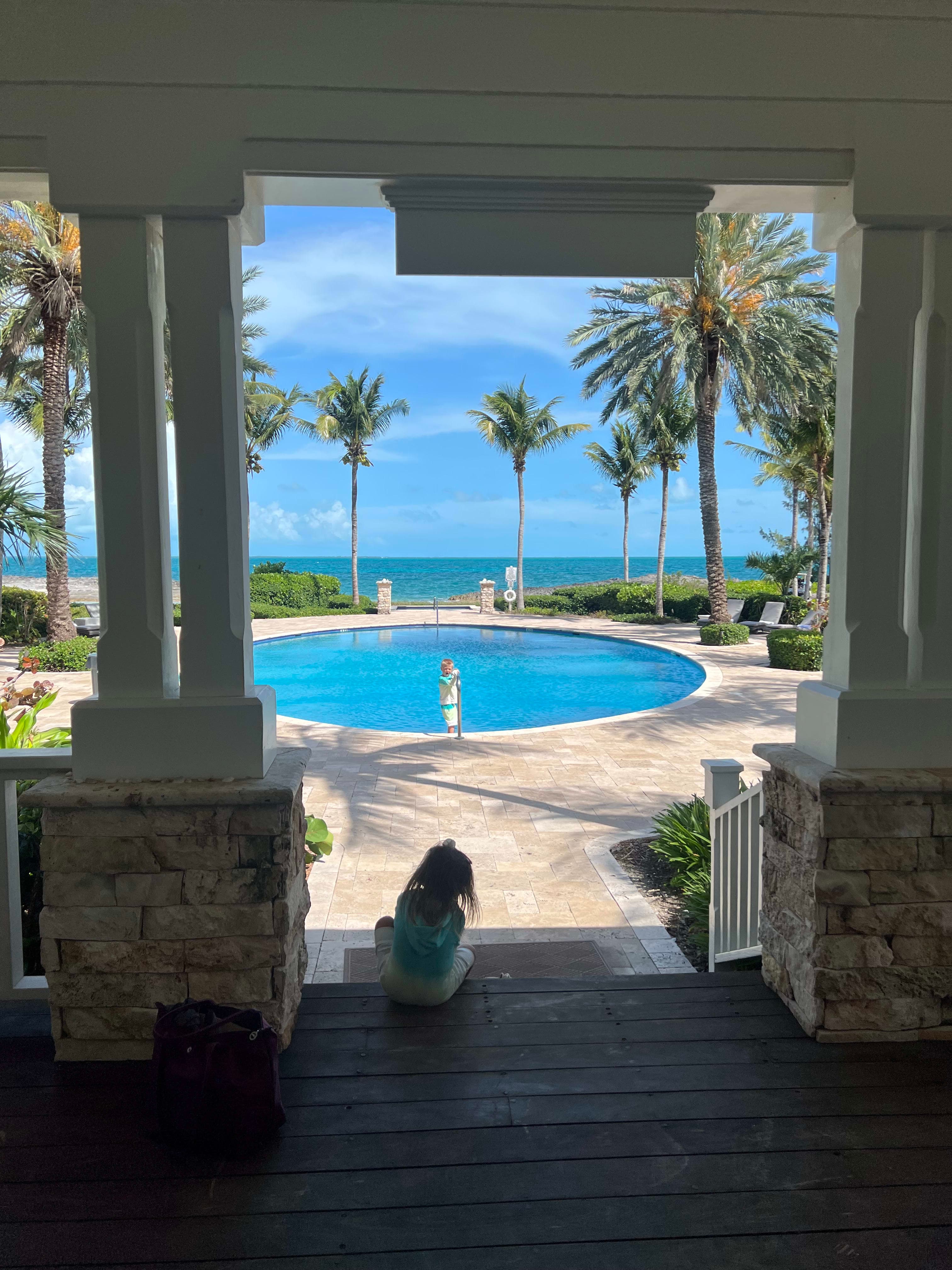 A child sitting on porch steps overlooking a round pool with palm trees and the beach in the background.