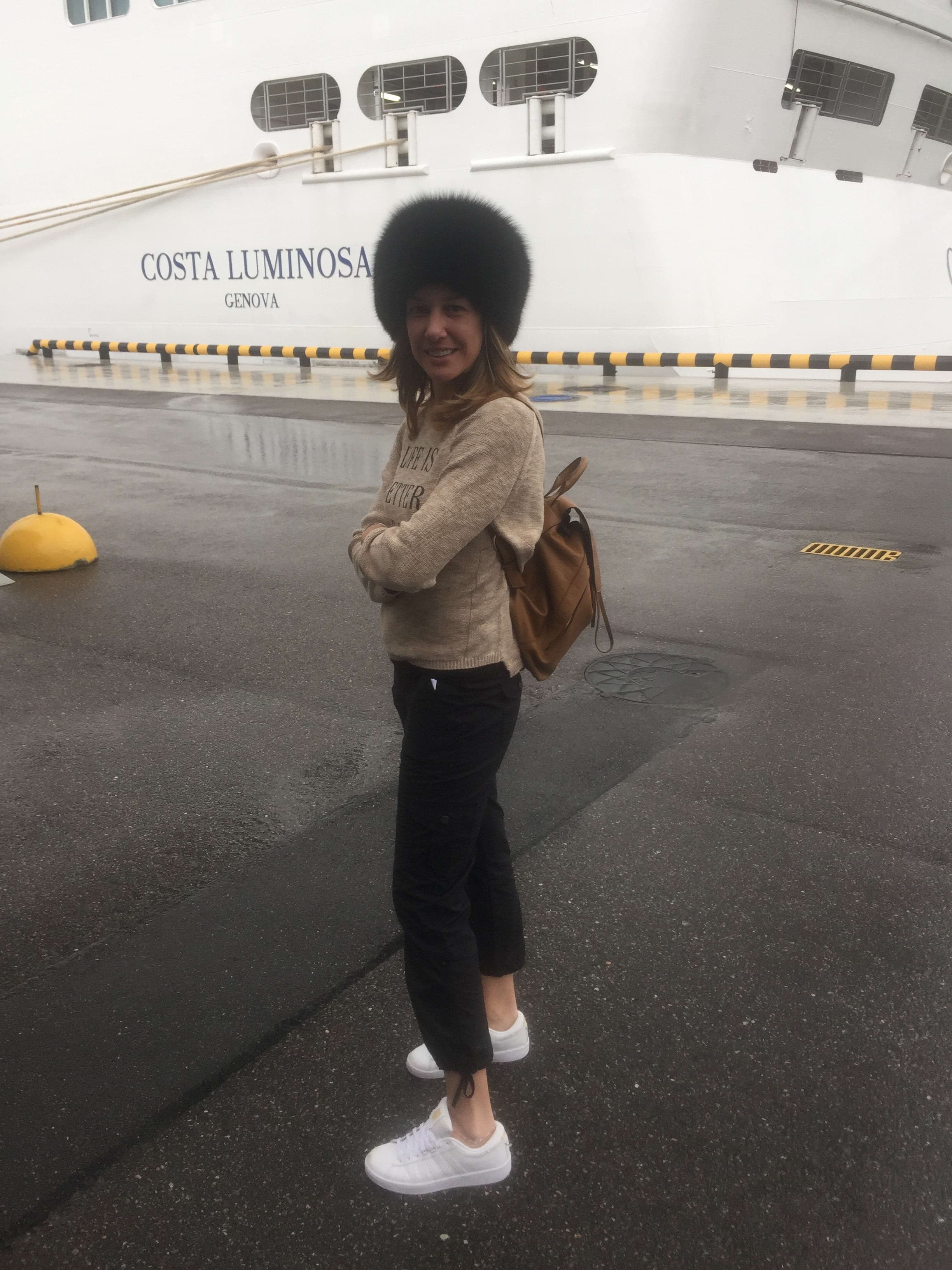 Debra wearing a large winter hat and standing on a dock in front of a white cruise ship