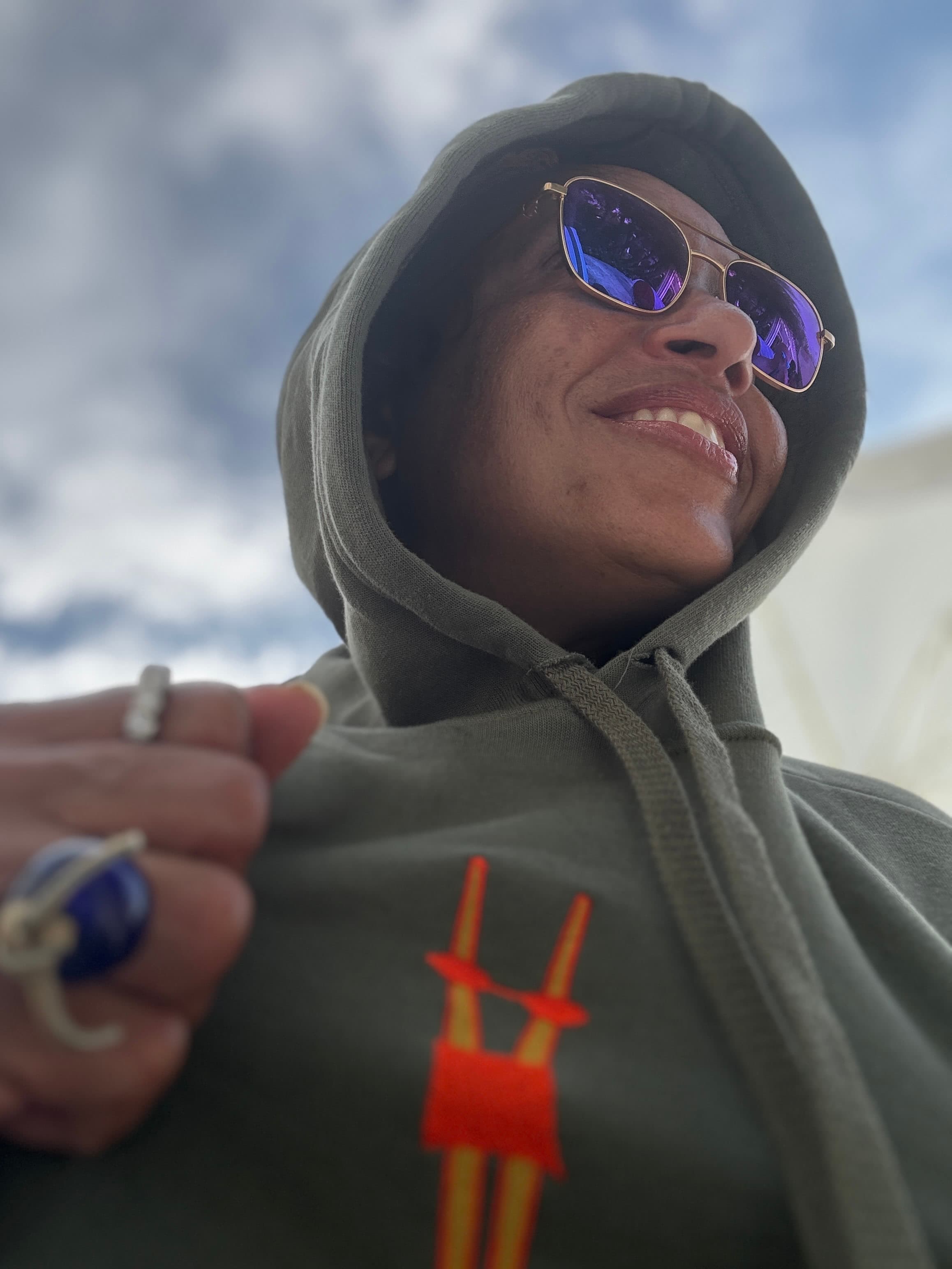 A photo taken from below of Peta smiling while wearing a hoodie and sunglasses on a cloudy day
