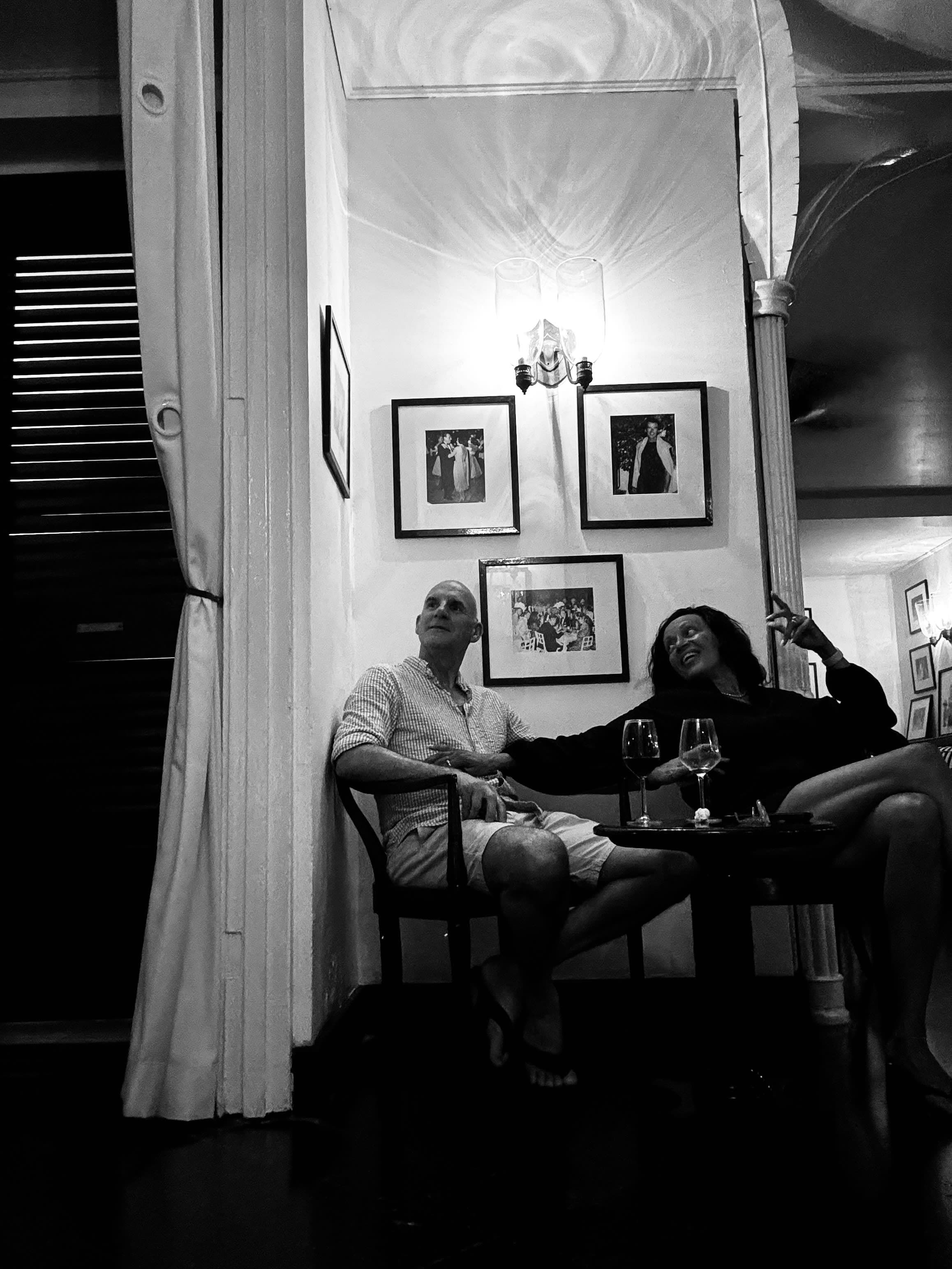 A greyscale photo of Peta with a male partner sitting on wooden chairs and having drinks