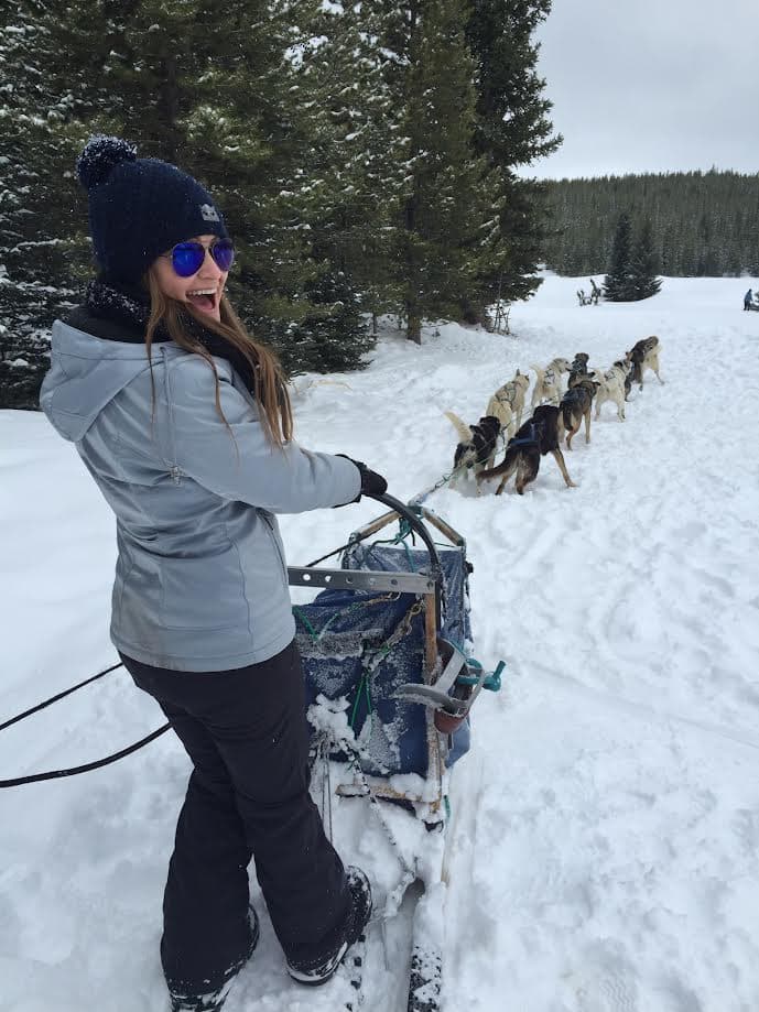 Brianna wearing winter gear standing on a sled pulled by sled dogs with surroundings covered by deep snow