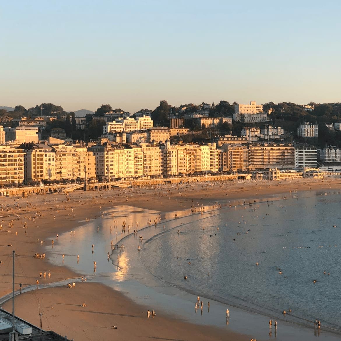 A beach shore at dusk with buildings in the distance and people walking on the beach.