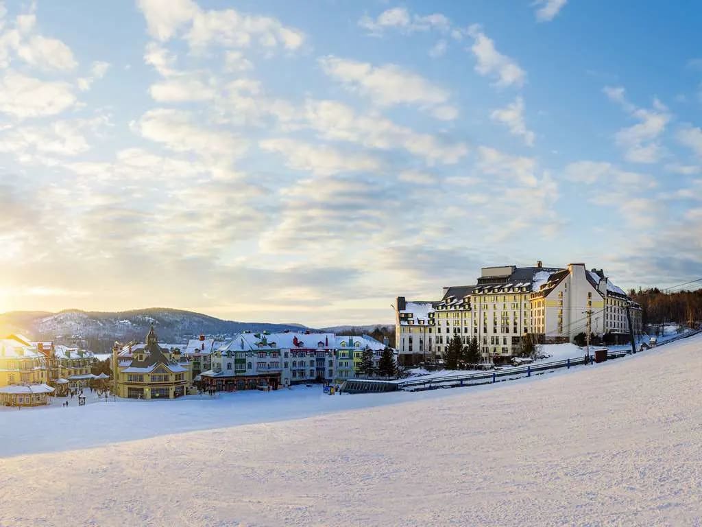 a white hotel in winter with ski slopes in the foreground and mountains in the background below a party cloudy blue sky
