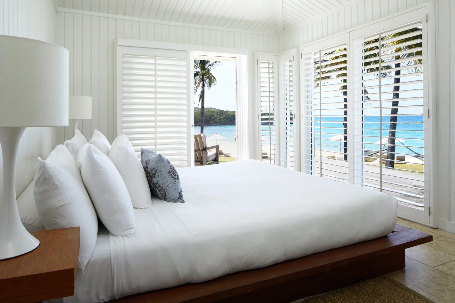 A white wood wall hotel room with white linens, dark wood bed frame and nightstand, with ocean outside.