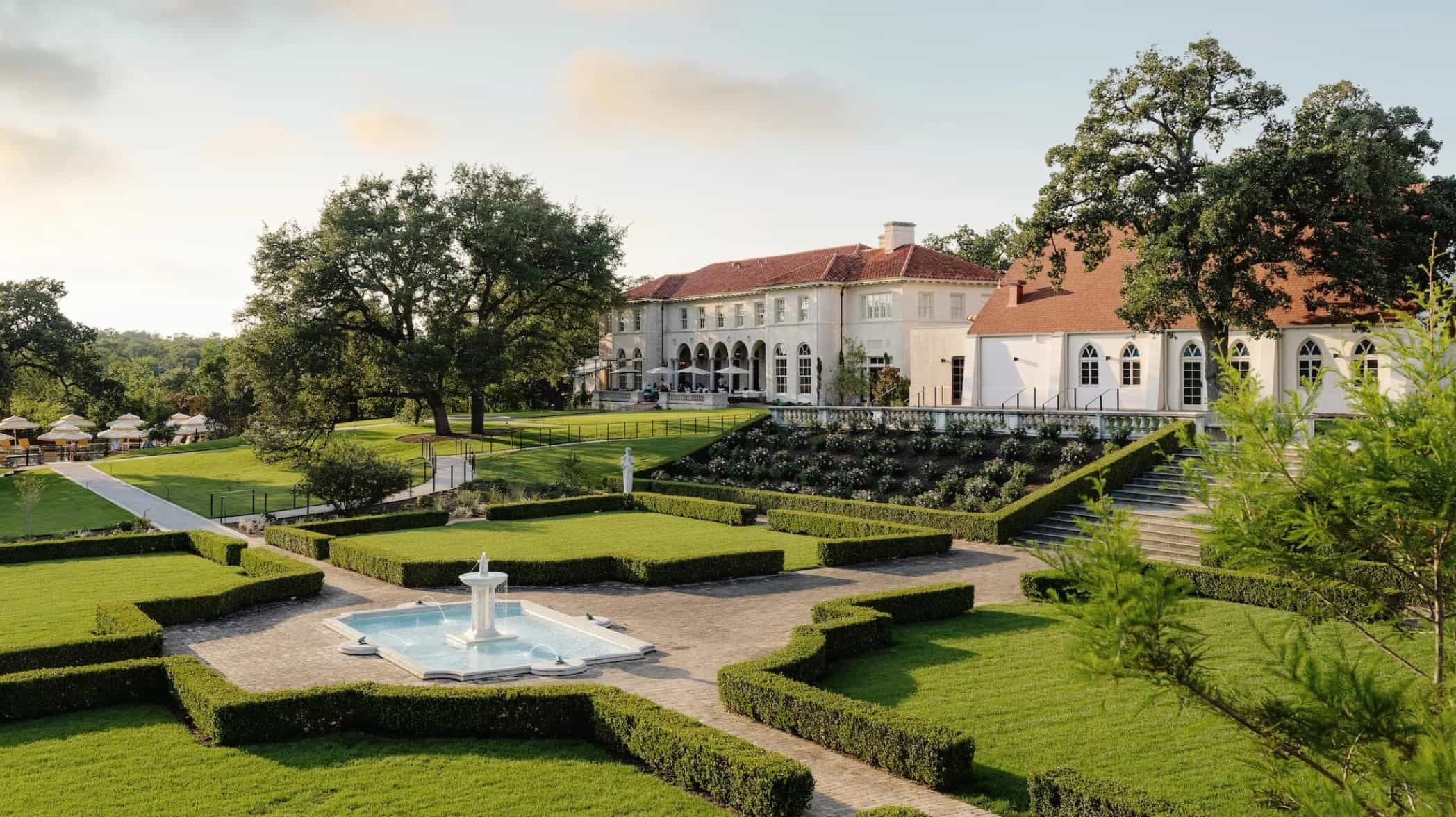 View of exapansive estate with green garden hedges, white fountain, and white estate building with tan roof in the background