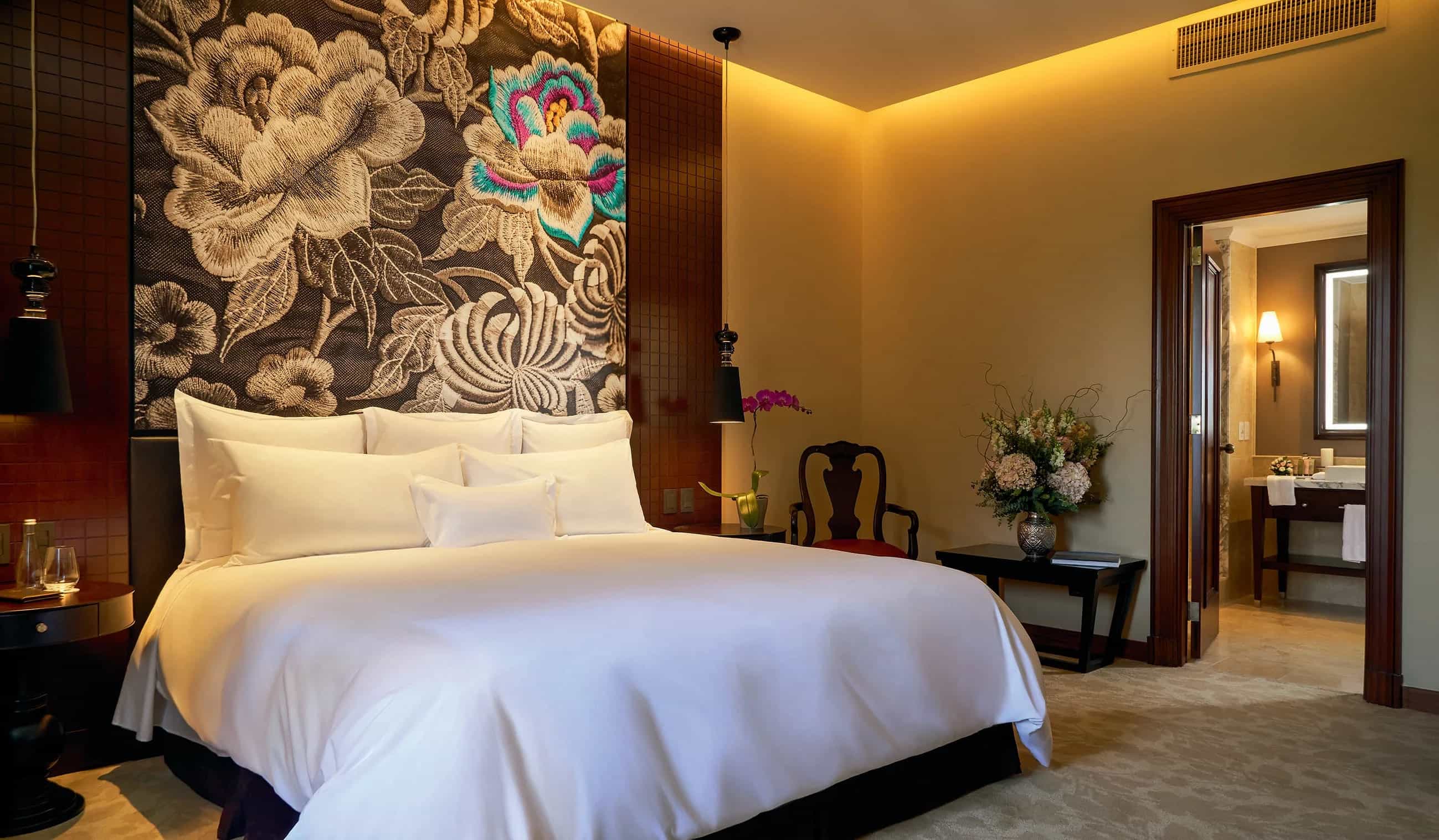 white linen covered bed with floral headboard up to the ceiling in a hotel room
