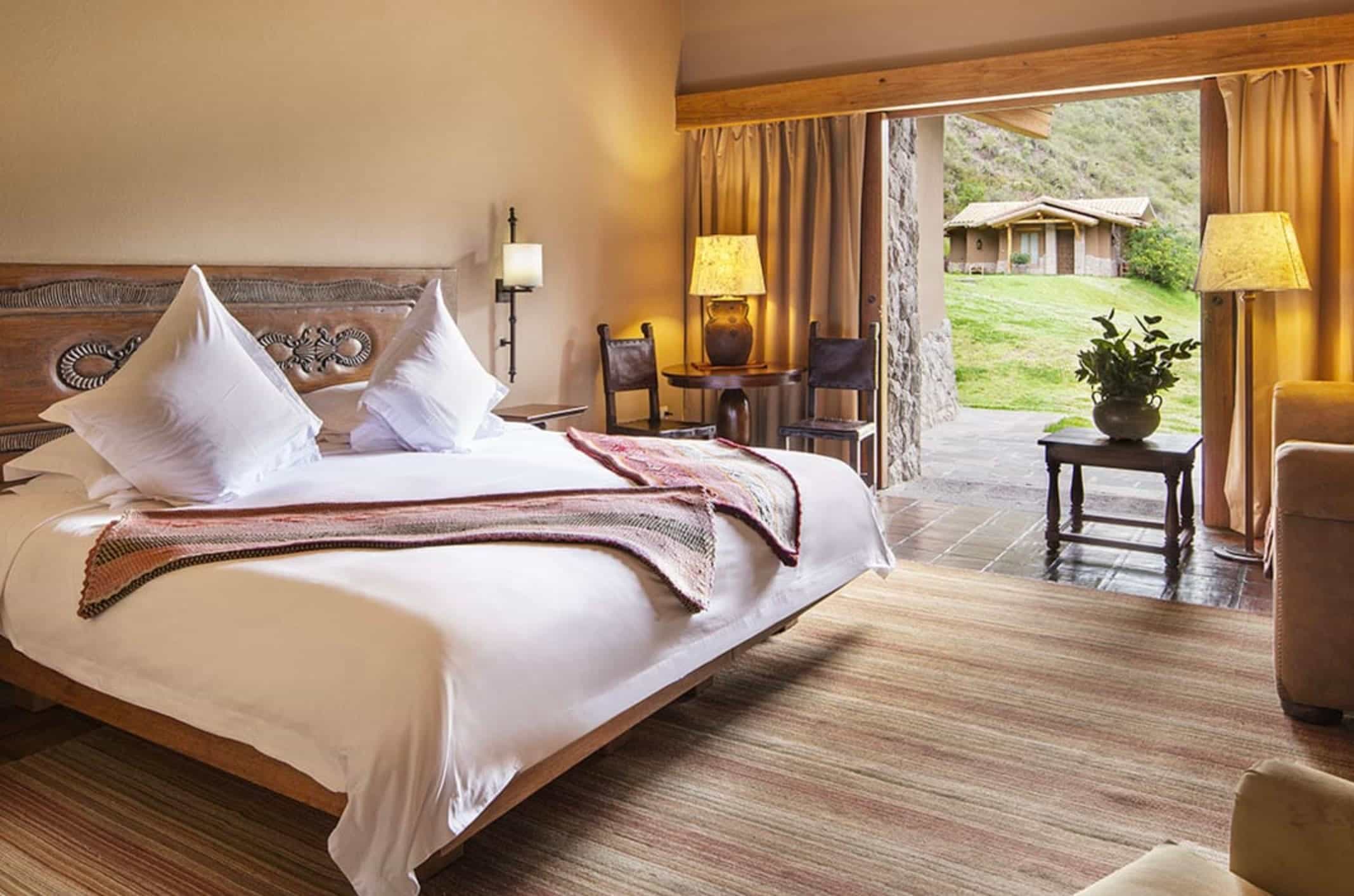 A rustic hotel room with white linen bed, carved wood headboard and open doors to a terrace with green hills