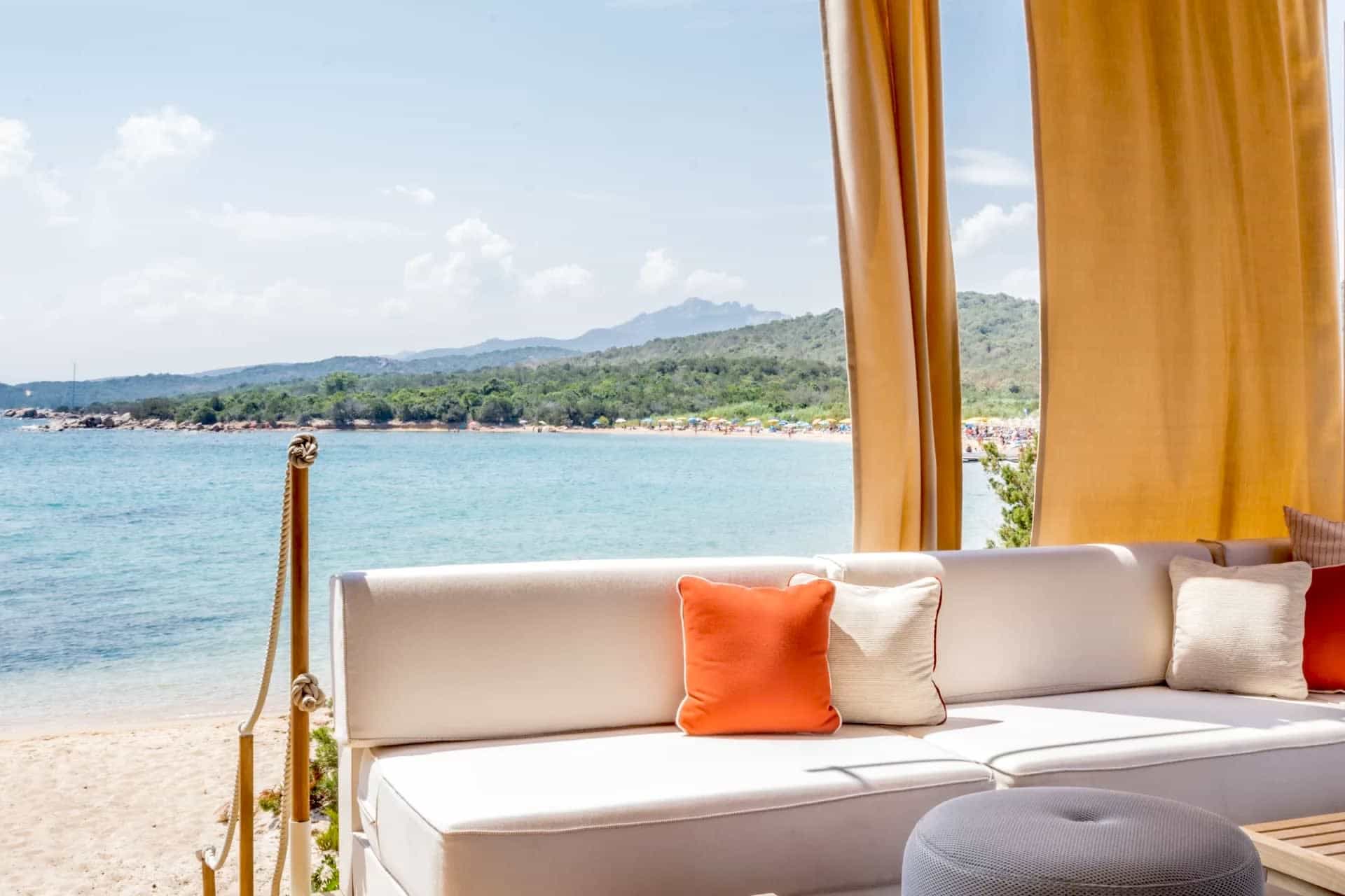 Insider's Guide to Sardinia White couch with gold curtains, and a beach and ocean behind