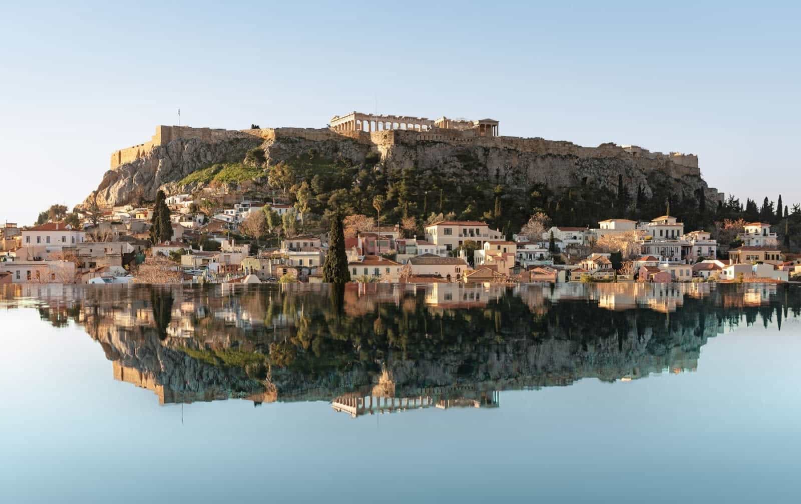 ancient ruins on top of a hill with buildings at the base all reflected in water during daytime