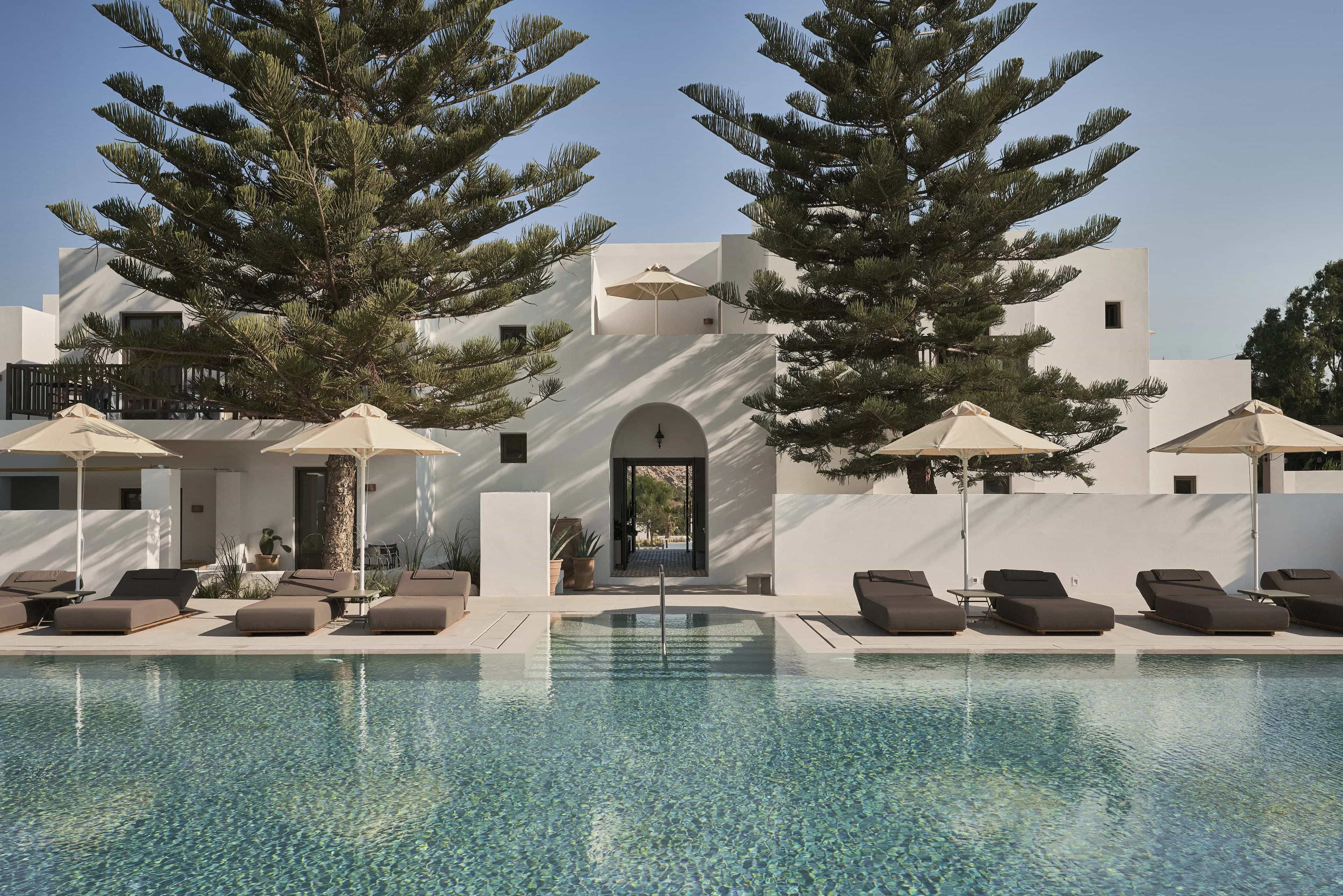 A pool with grey sun loungers, white umbrellas in front of a white stucco building and tall pine trees during day