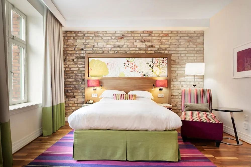 bright hotel room with multi colored purple and pink carpet, green bed skirt, and floral headboard with brick wall.