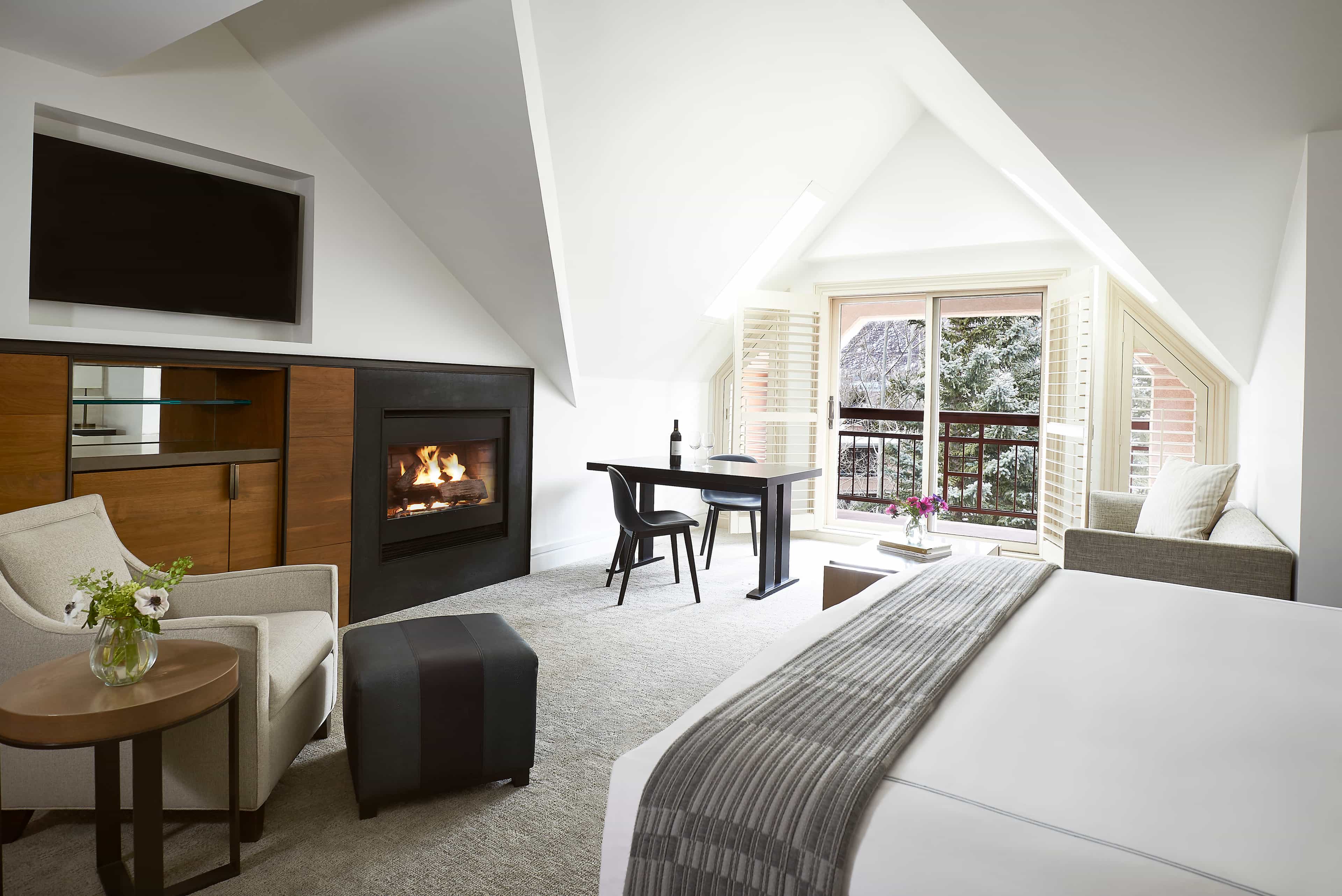Bright hotel room with sloped ceilings, a fireplace, and modern furnishings opening onto a balcony with mountain views.