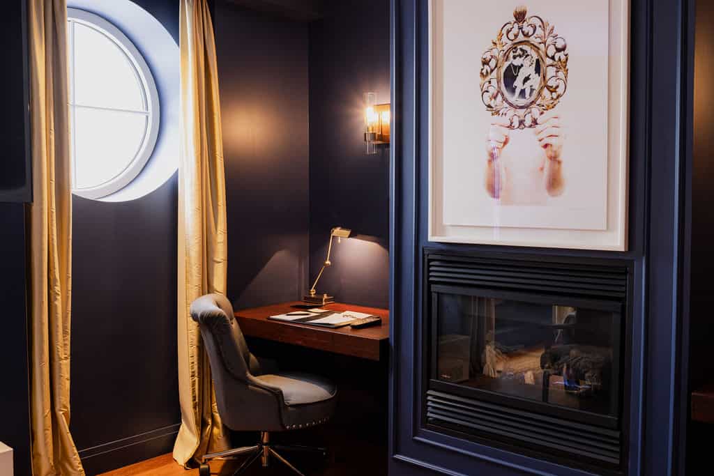 Cozy corner with a writing desk, round window, and a fireplace set into a navy wall topped with modern artwork.