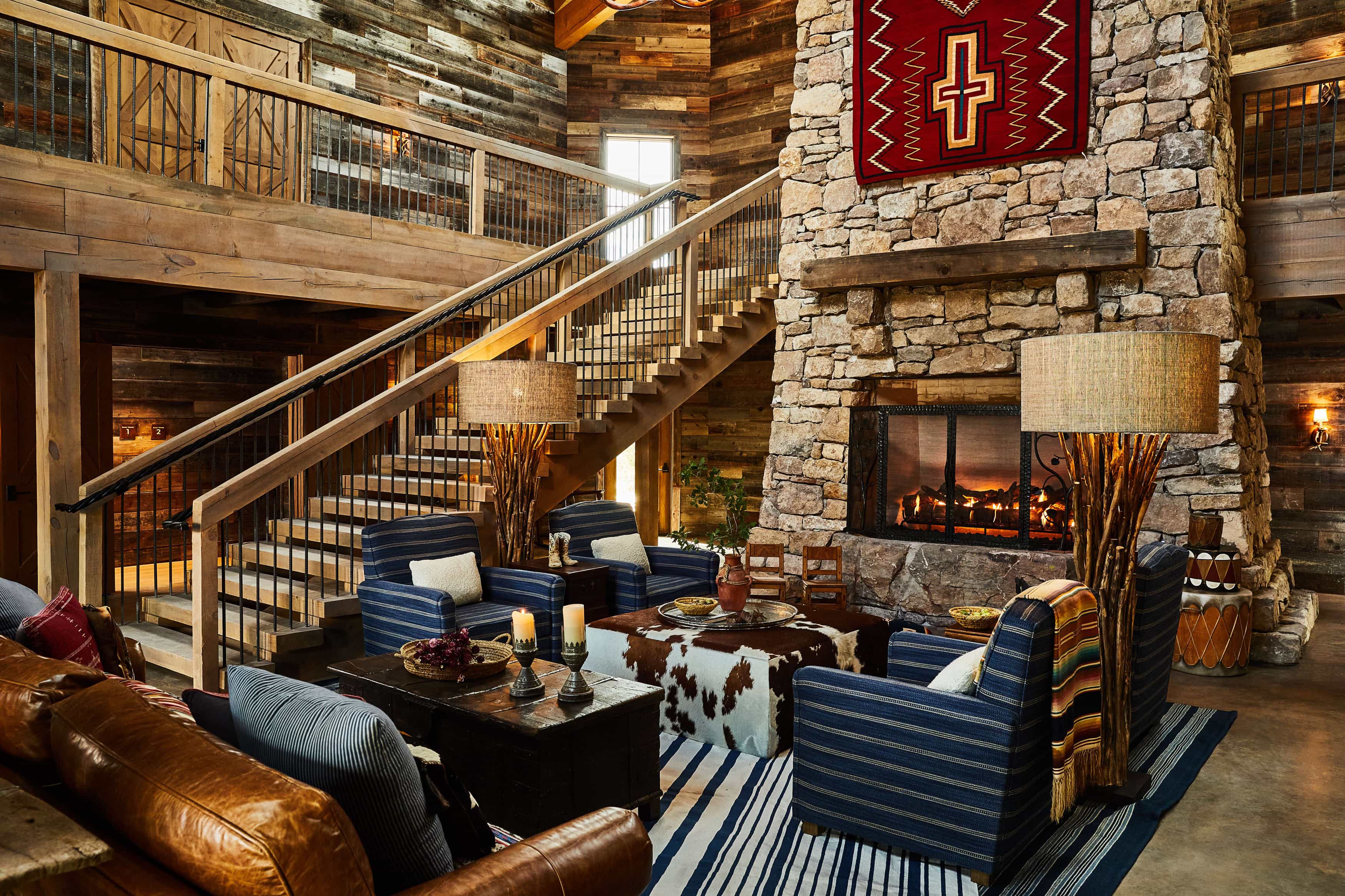 Rustic lodge living room with a stone fireplace, leather and striped armchairs, wooden beams, and a staircase leading to a loft.