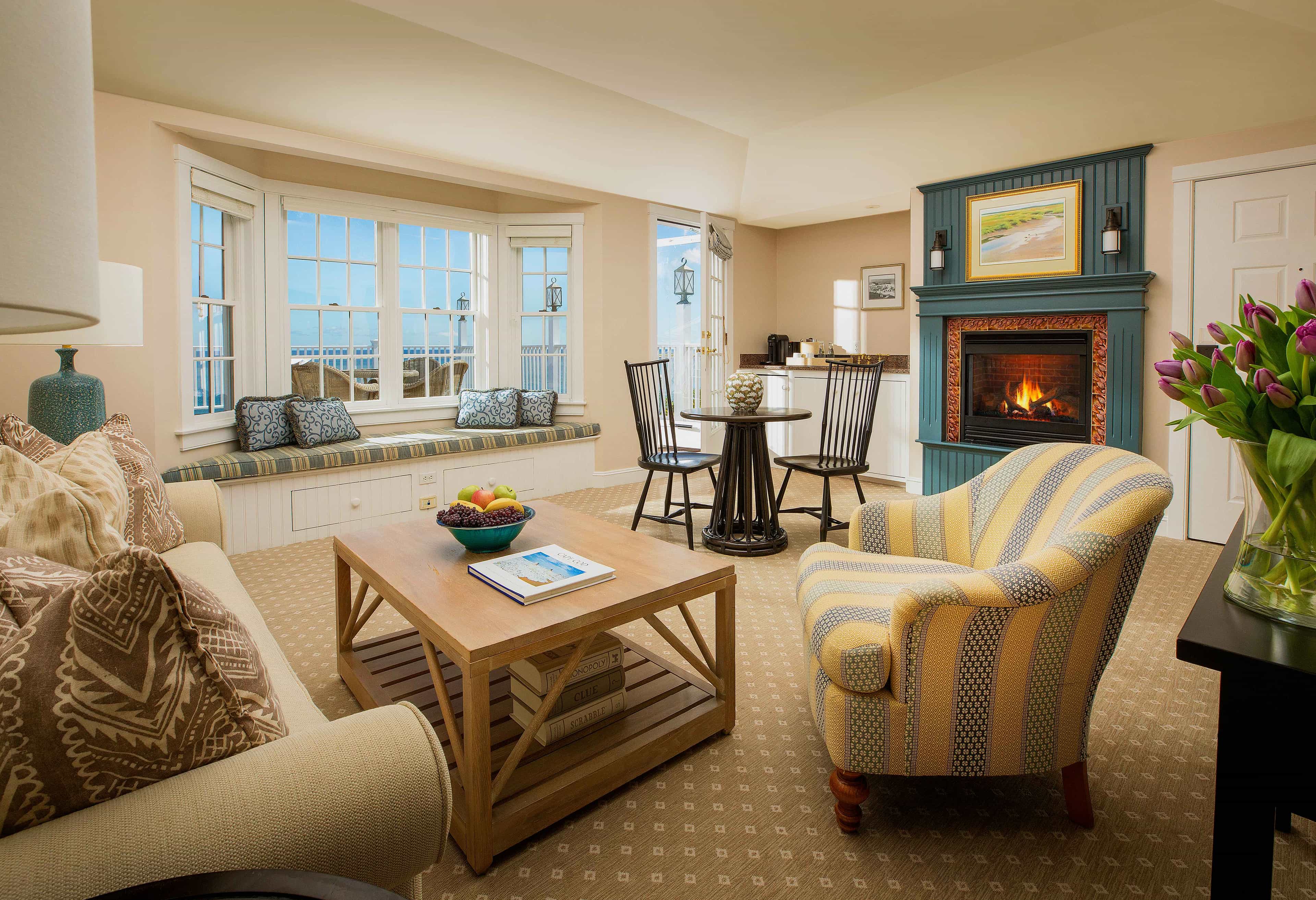 Bright coastal-style living room with patterned armchairs, a teal fireplace, bay window seating, and views of the ocean.