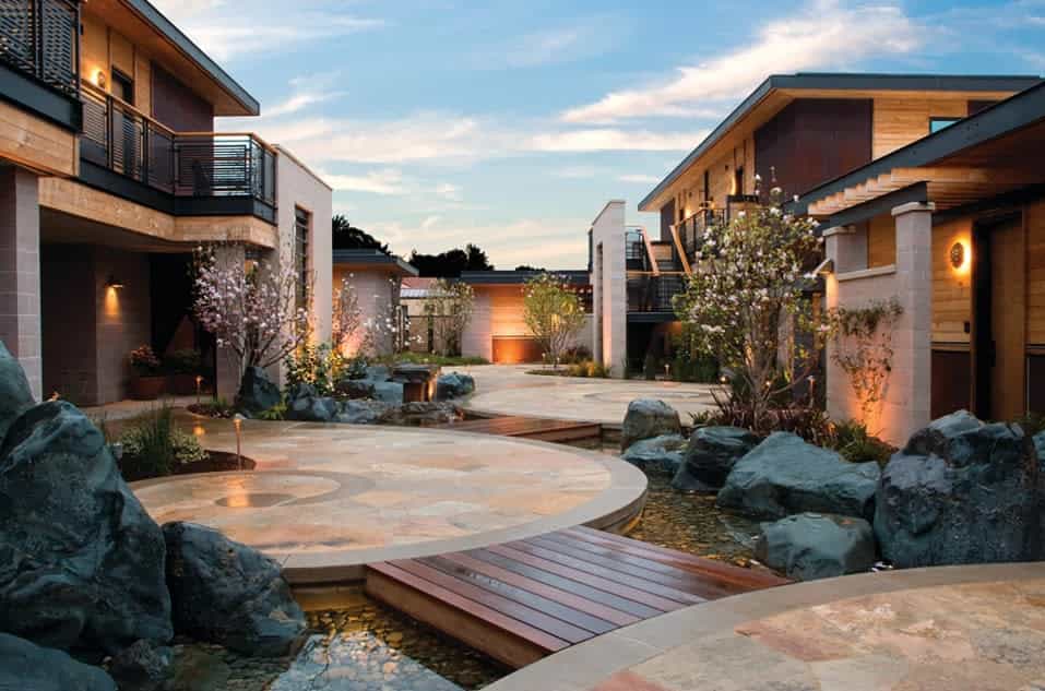 Modern courtyard with stone paths, water features, blossoming trees, and contemporary buildings at sunset.