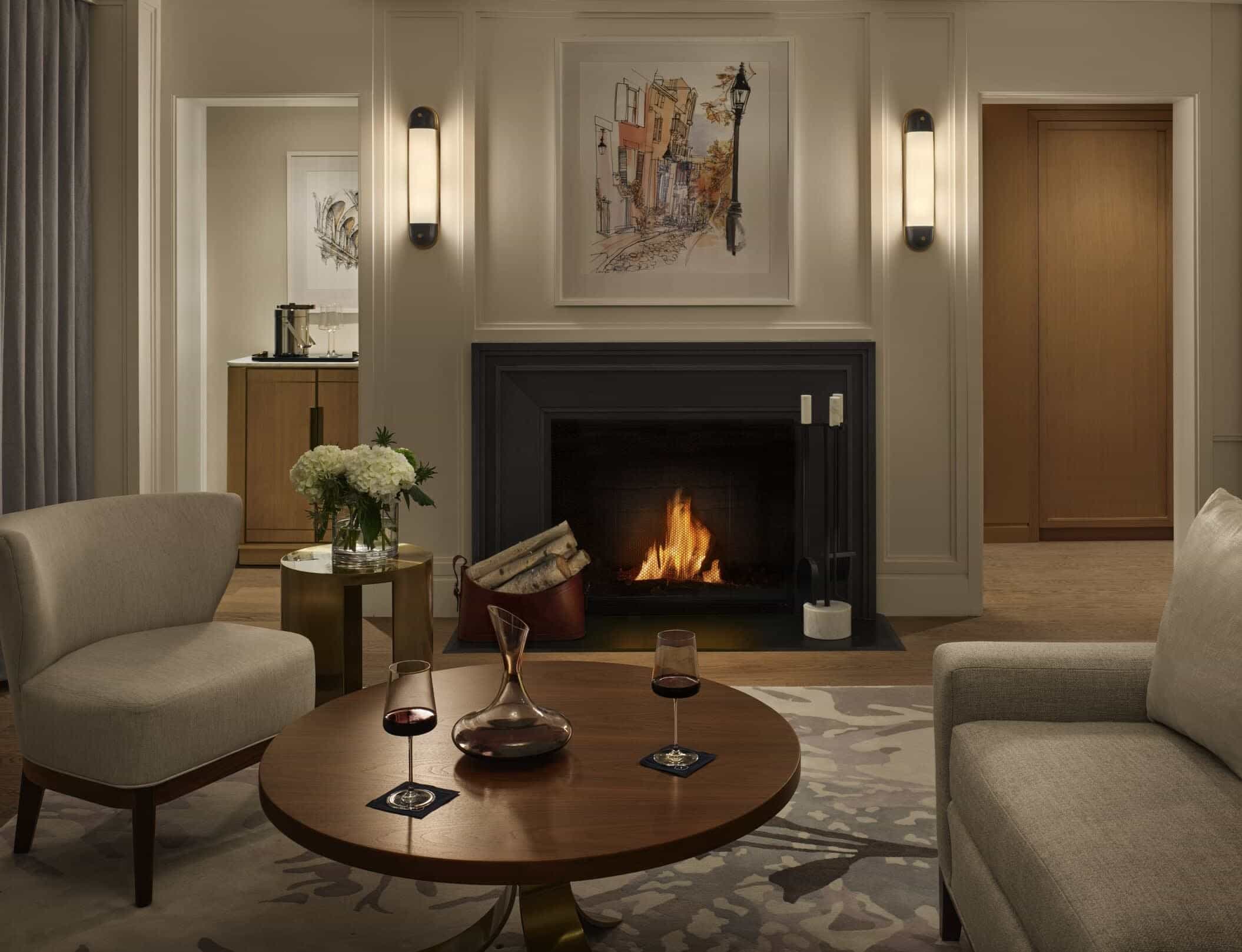 Elegant hotel suite with a cozy fireplace, modern armchairs, fresh flowers, and wine glasses on a wooden coffee table.