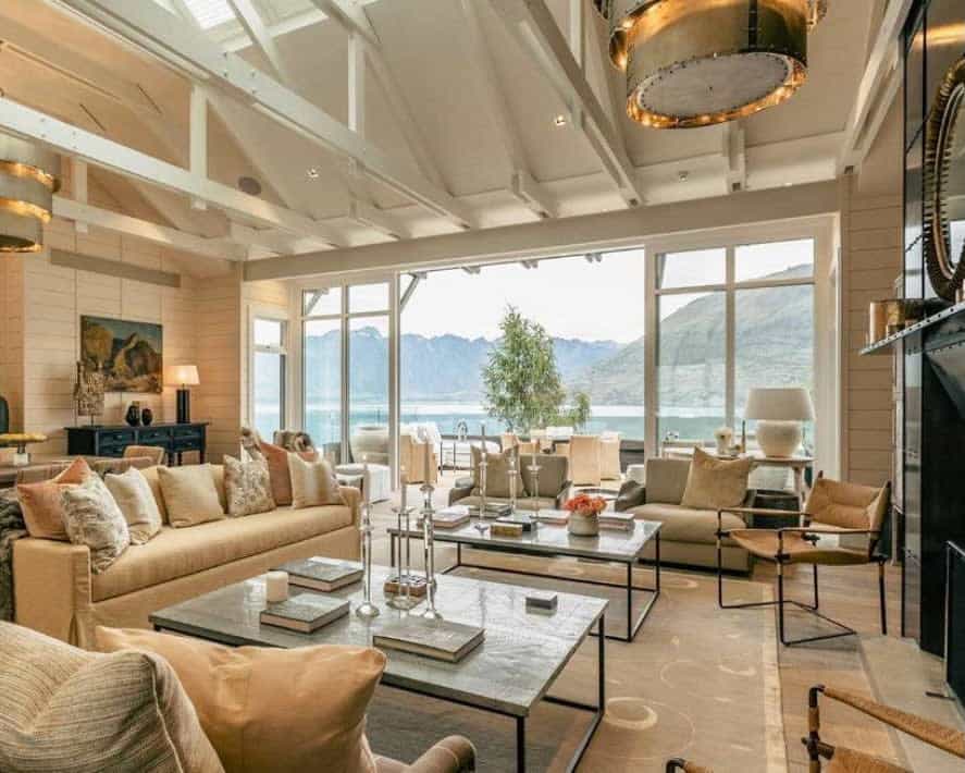 contemporary villa living room with cream colored furniture, glass tables, wood beam ceiling, and mountains and lake outside