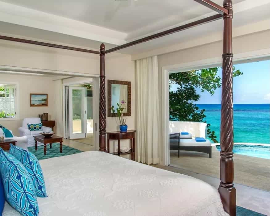 A white hotel room with dark wood four poster bed facing out an open wall to oceanfront infinity pool during day