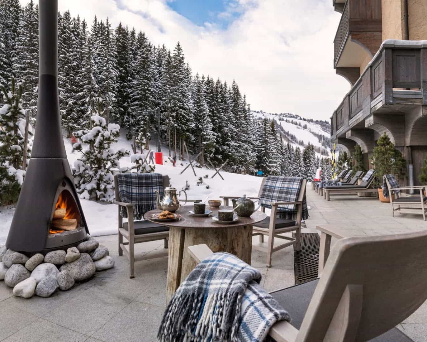 Outdoor terrace with plaid blankets covering chairs next to a fireplace and slow covered trees beyond during day
