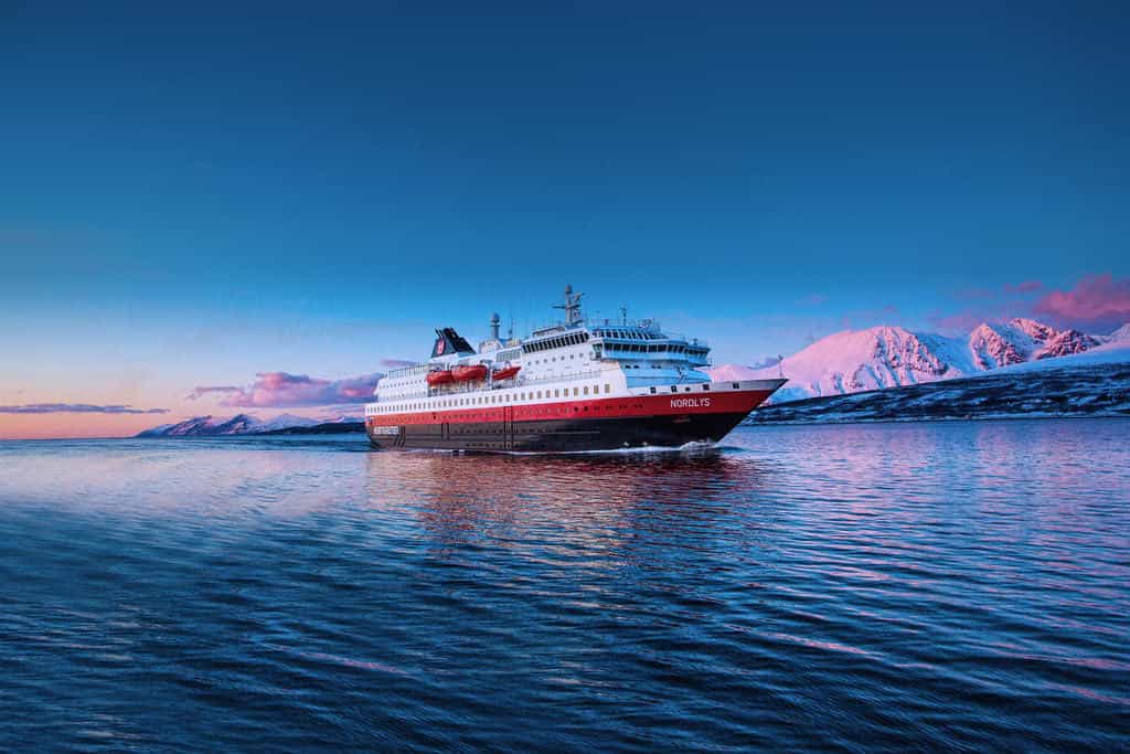 A white, red, and black ship at sea with snow covered mountains at dusk in the background